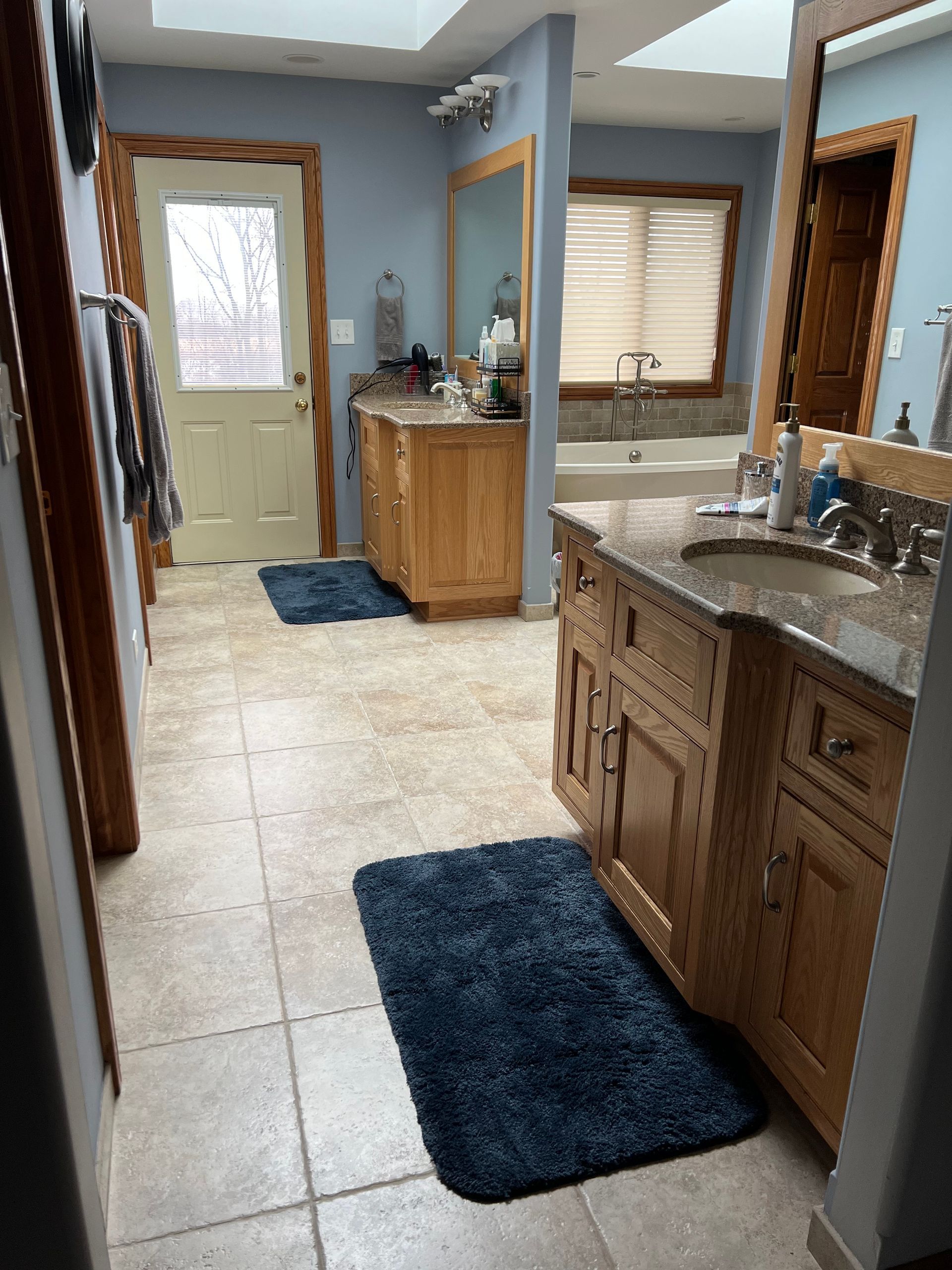 Bathroom with double sink vanity, mirrors, windows, and blue rugs on a tiled floor.