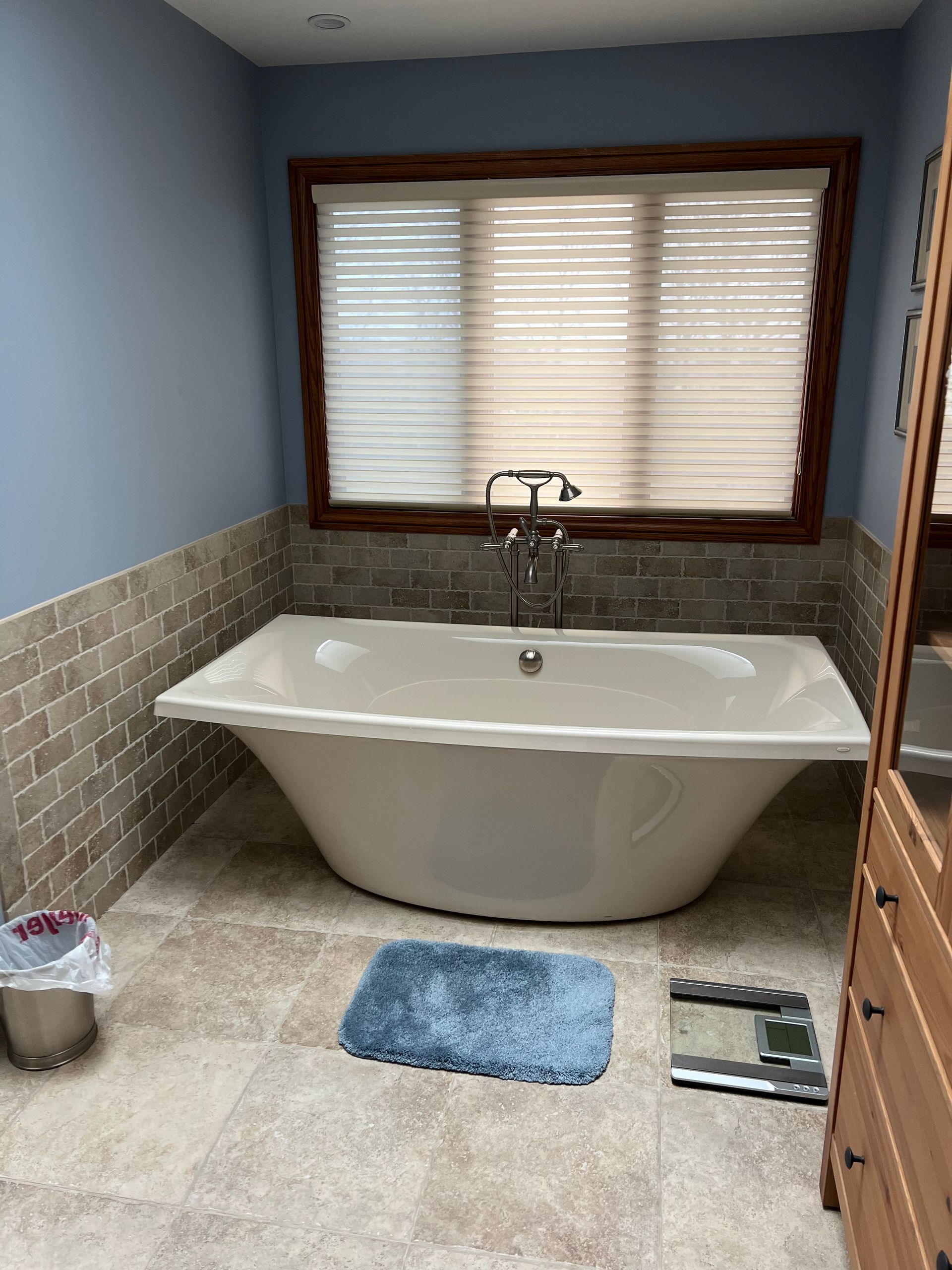 Bathroom with a freestanding white bathtub, blue walls, tiled wainscoting, window blinds, and bath mats.