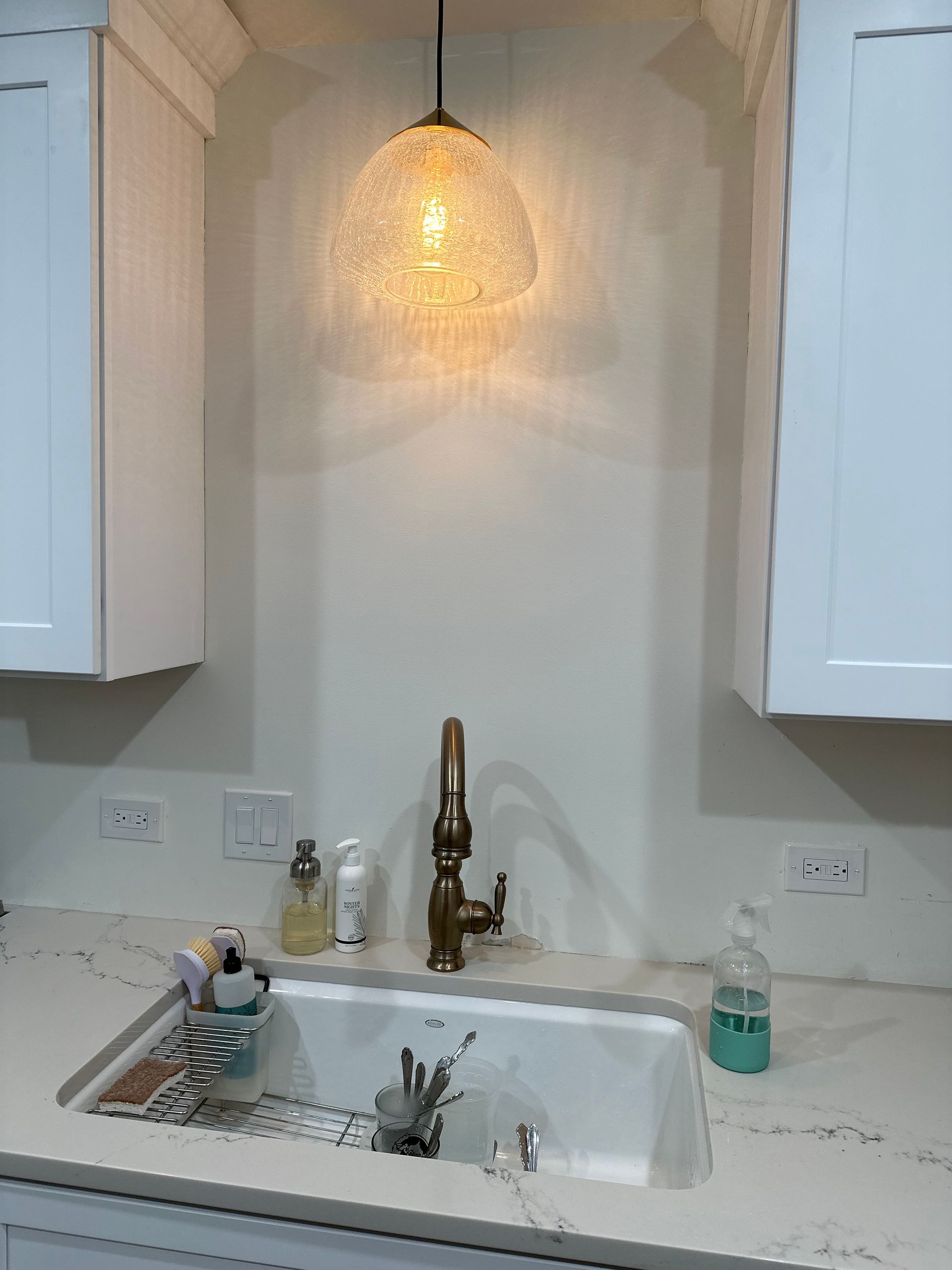 White kitchen sink alcove with brass faucet, pendant light, and cleaning supplies on the counter
