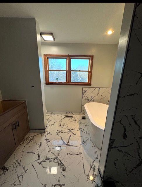 Small bathroom with marble-look tile floor, window, vanity, and white tub under warm lights