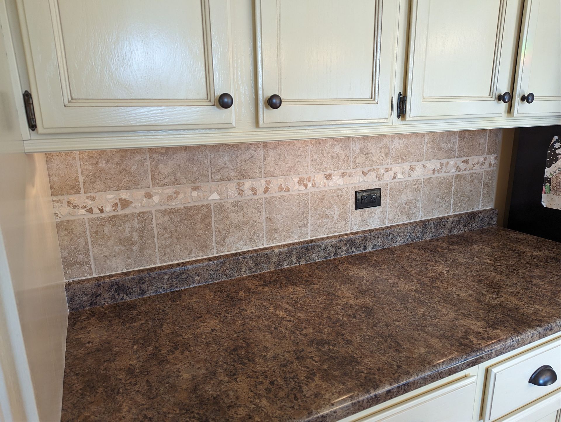 Kitchen countertop with brown speckled surface, beige tiled backsplash, and cream cabinets above
