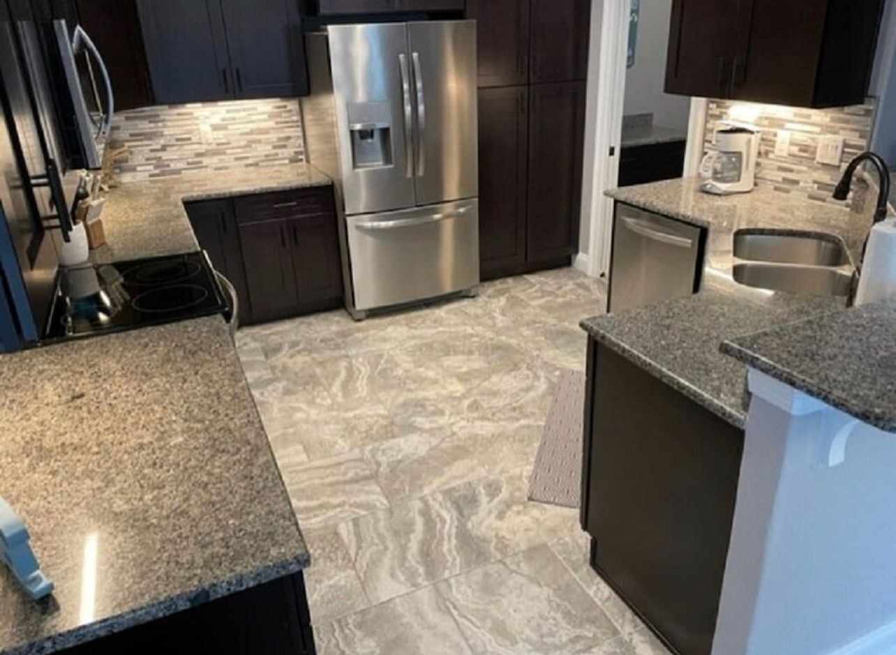 Modern kitchen with stainless steel fridge, dark cabinets, speckled counters, and light tiled floor.