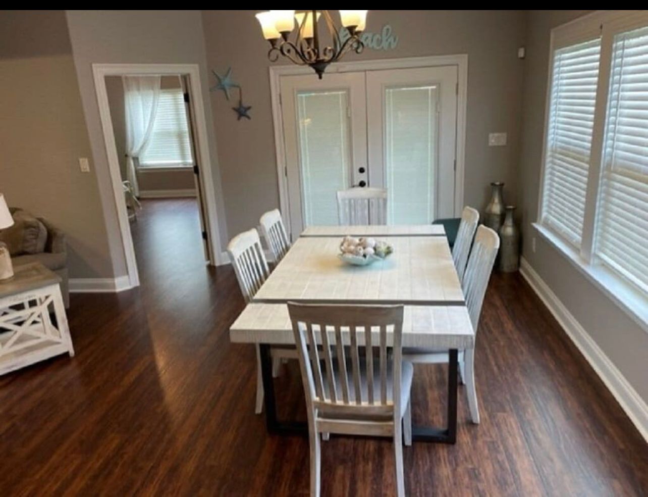 Dining room with white table and chairs, dark wood floor, and large windows with blinds