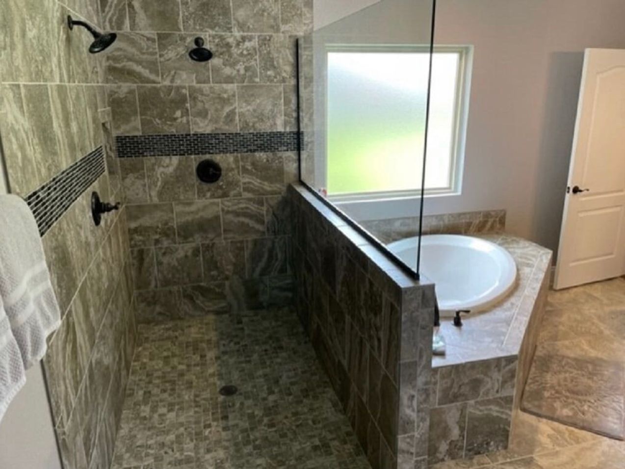 Walk-in shower with stone tile, glass partition, and a soaking tub by a frosted window
