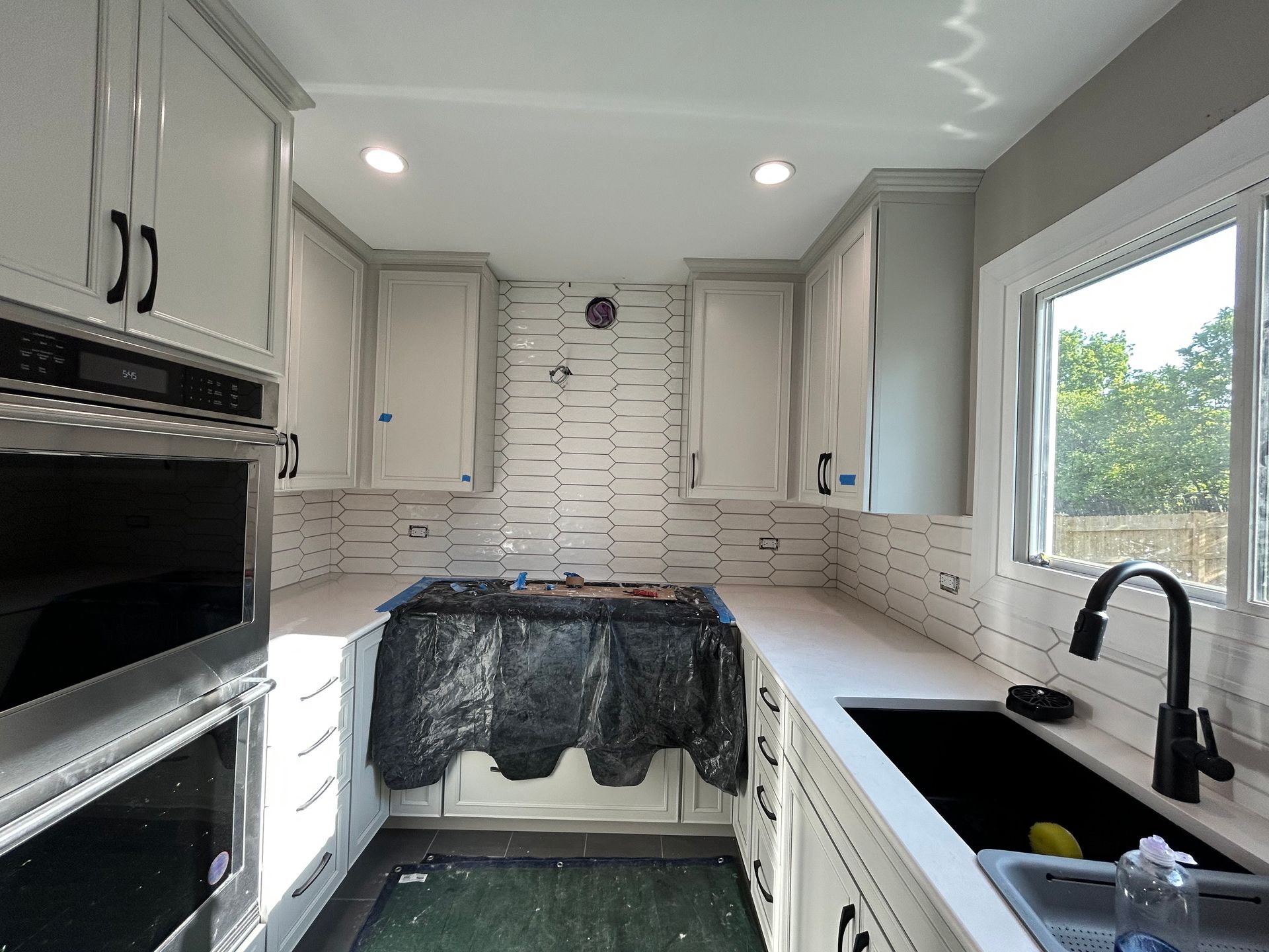 Kitchen under renovation with white cabinets, black sink, and covered island in a bright room