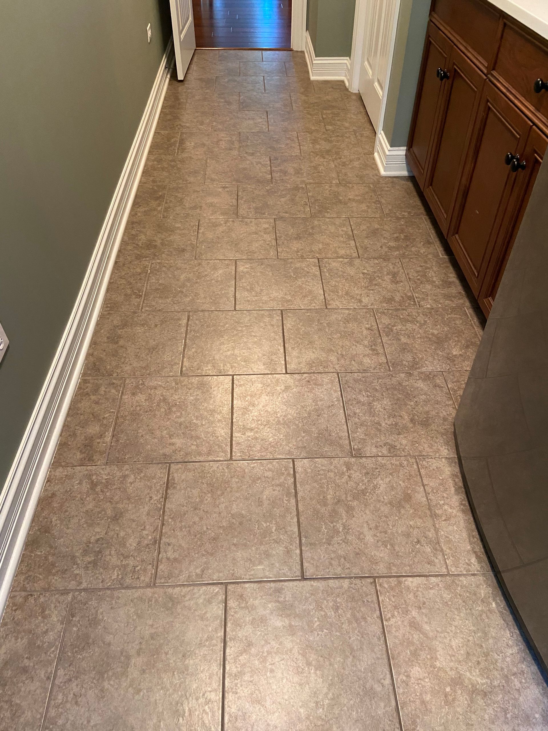Narrow hallway with tan tile floor, white baseboards, and wooden cabinets on the right.