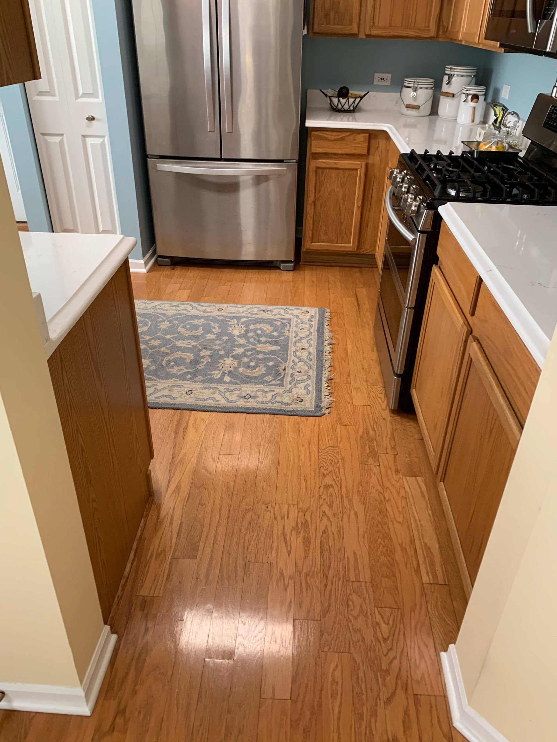 Kitchen with stainless steel fridge, wood cabinets, black stove, and a small rug on the hardwood floor.