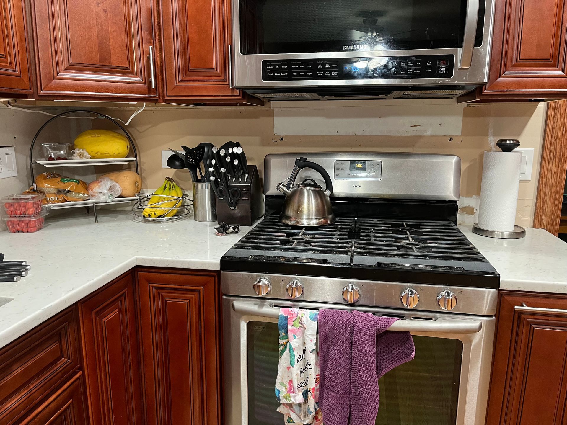 Kitchen with stainless steel stove and microwave, kettle on burner, wooden cabinets, and colorful towels on oven handle