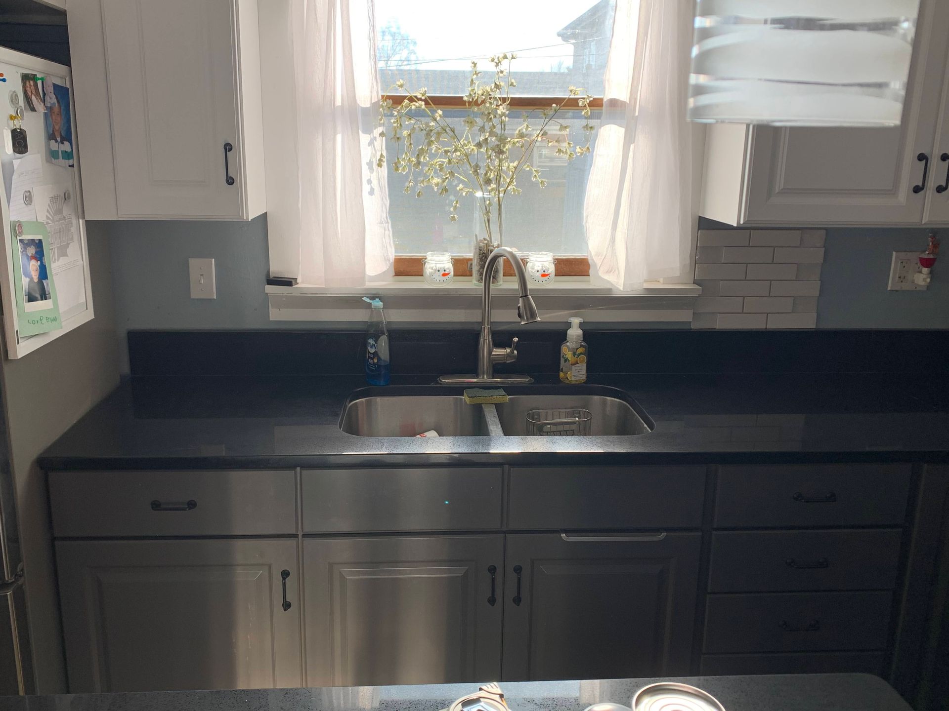 Kitchen sink under a window with dark cabinets, black countertop, and a vase of yellow flowers.