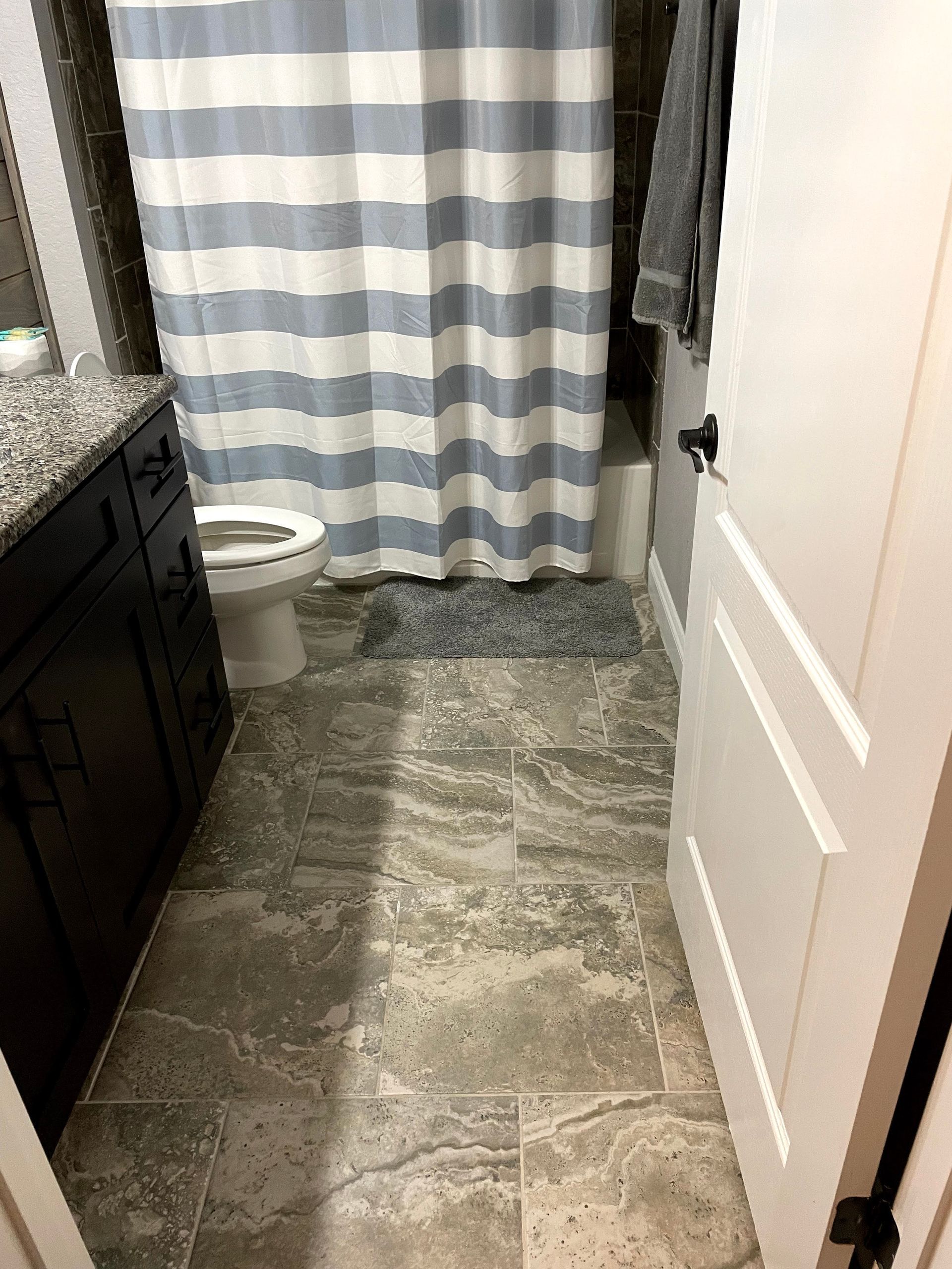 Narrow bathroom with tiled floor, striped shower curtain, toilet, sink, and open white door.