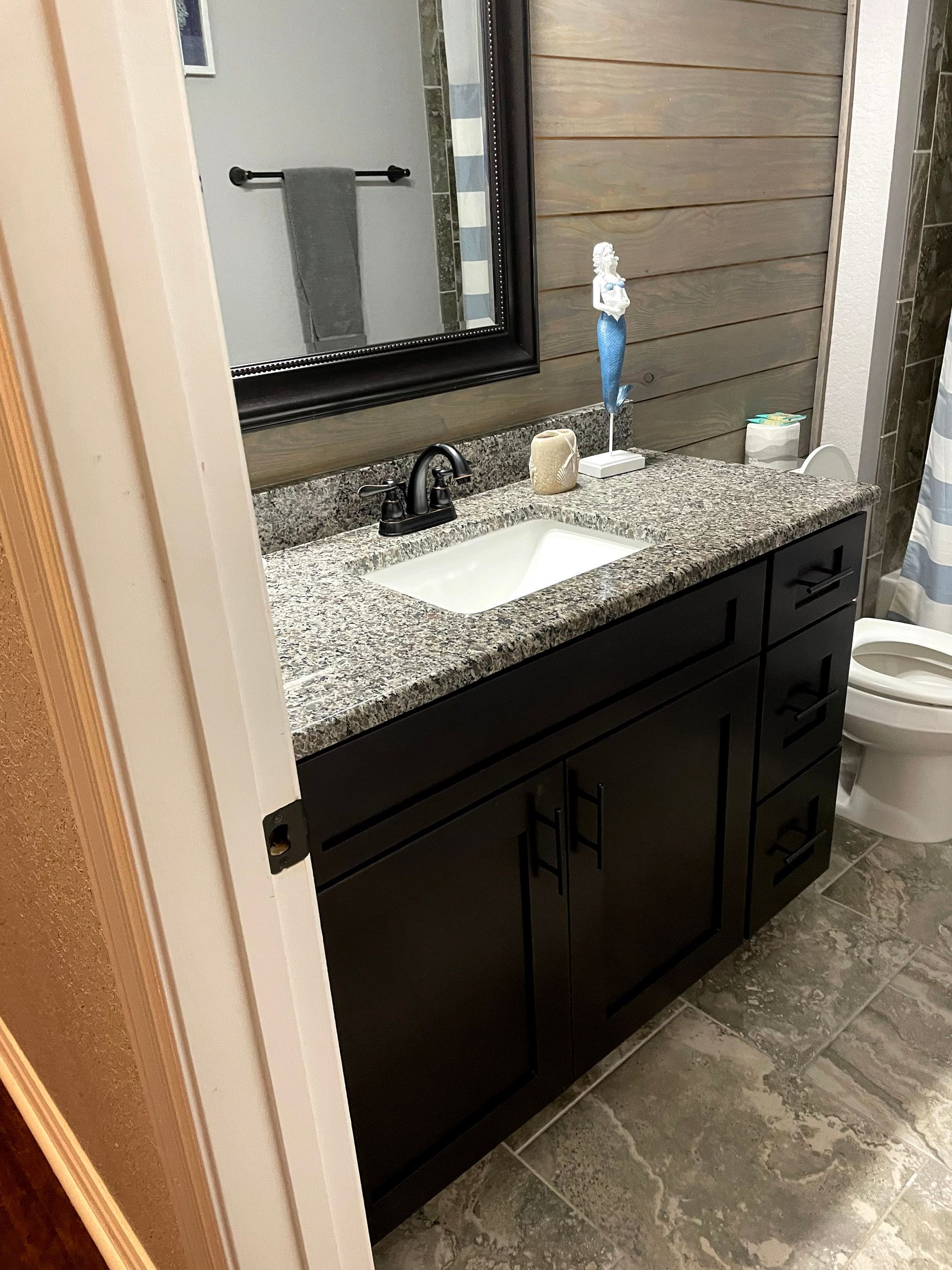 Bathroom vanity with dark cabinets, speckled countertop, sink, and large mirror beside a shower.