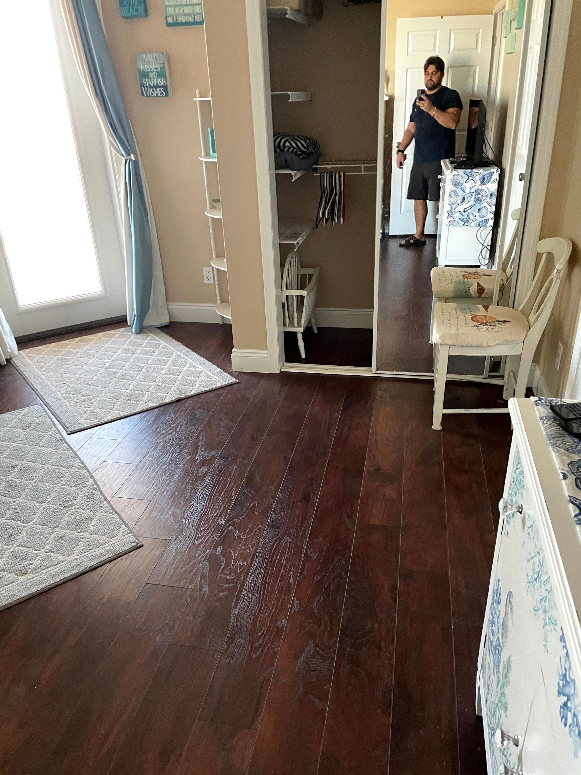 Mirror view of a living room with dark wood floor, rugs, chair, and a person taking the photo
