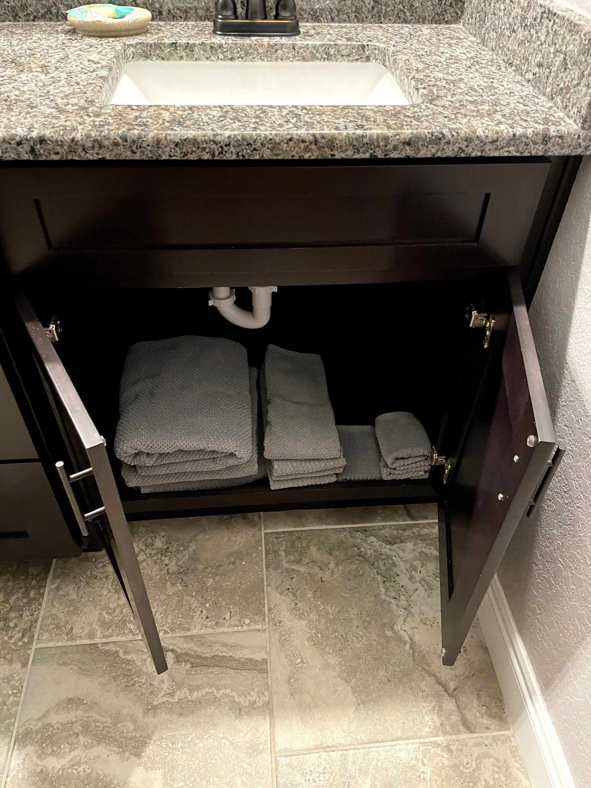 Open bathroom vanity cabinet with towels and plumbing visible under a granite sink.