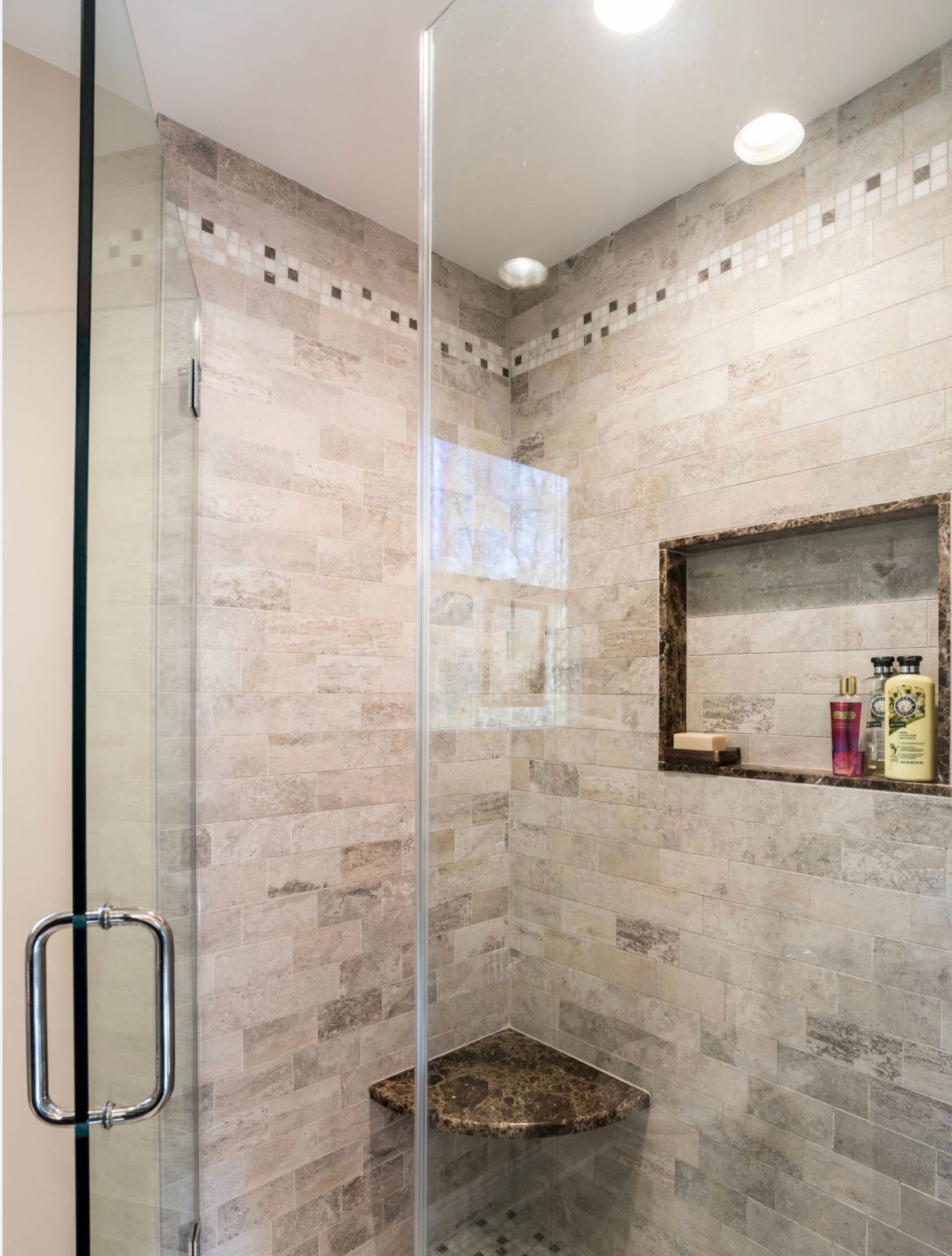 Glass shower with beige tiled walls, built-in corner shelf, and small wall niche with toiletries.