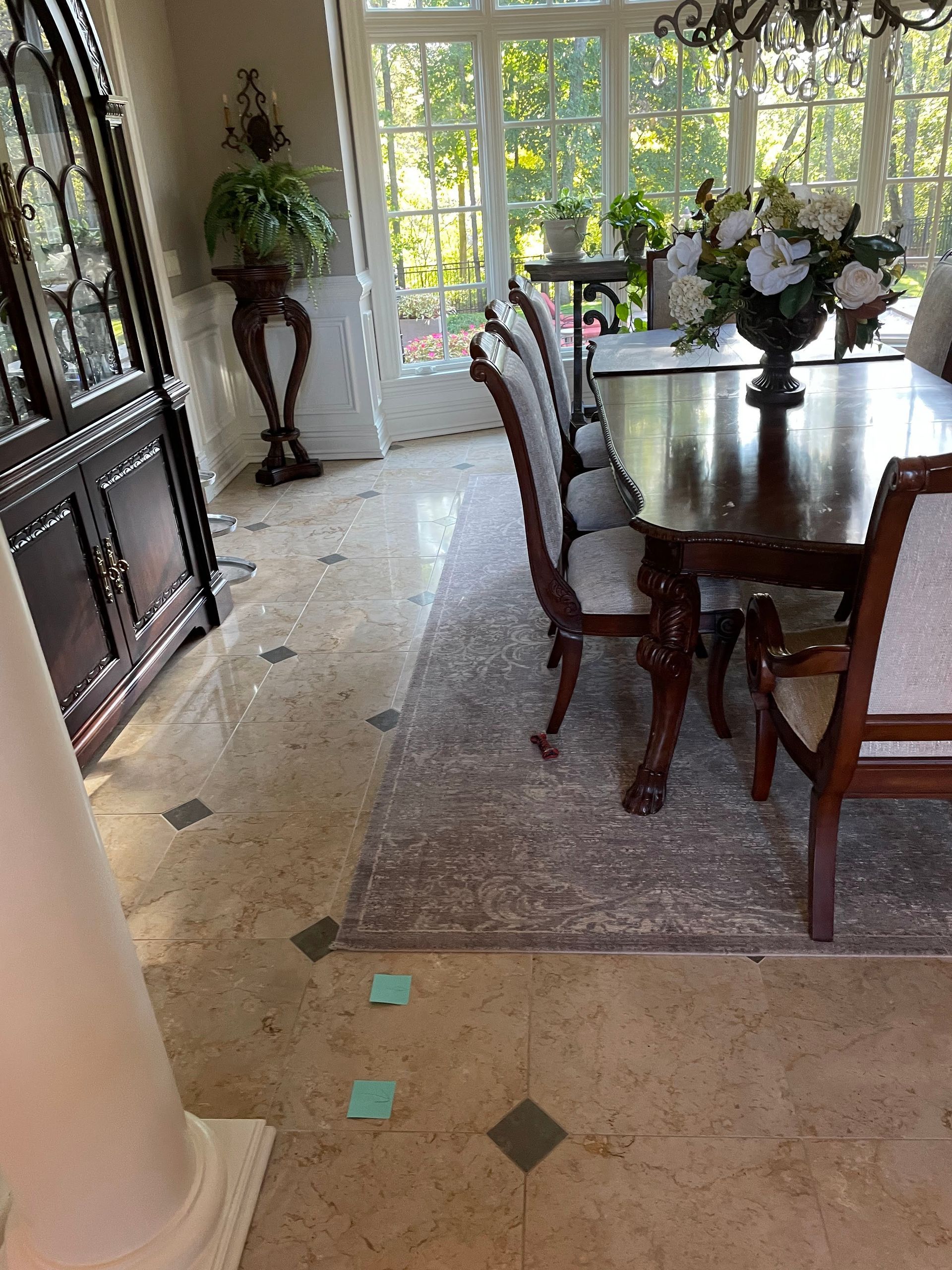 Elegant dining room with wooden table, chairs, floral centerpiece, patterned rug, and bright windows overlooking greenery