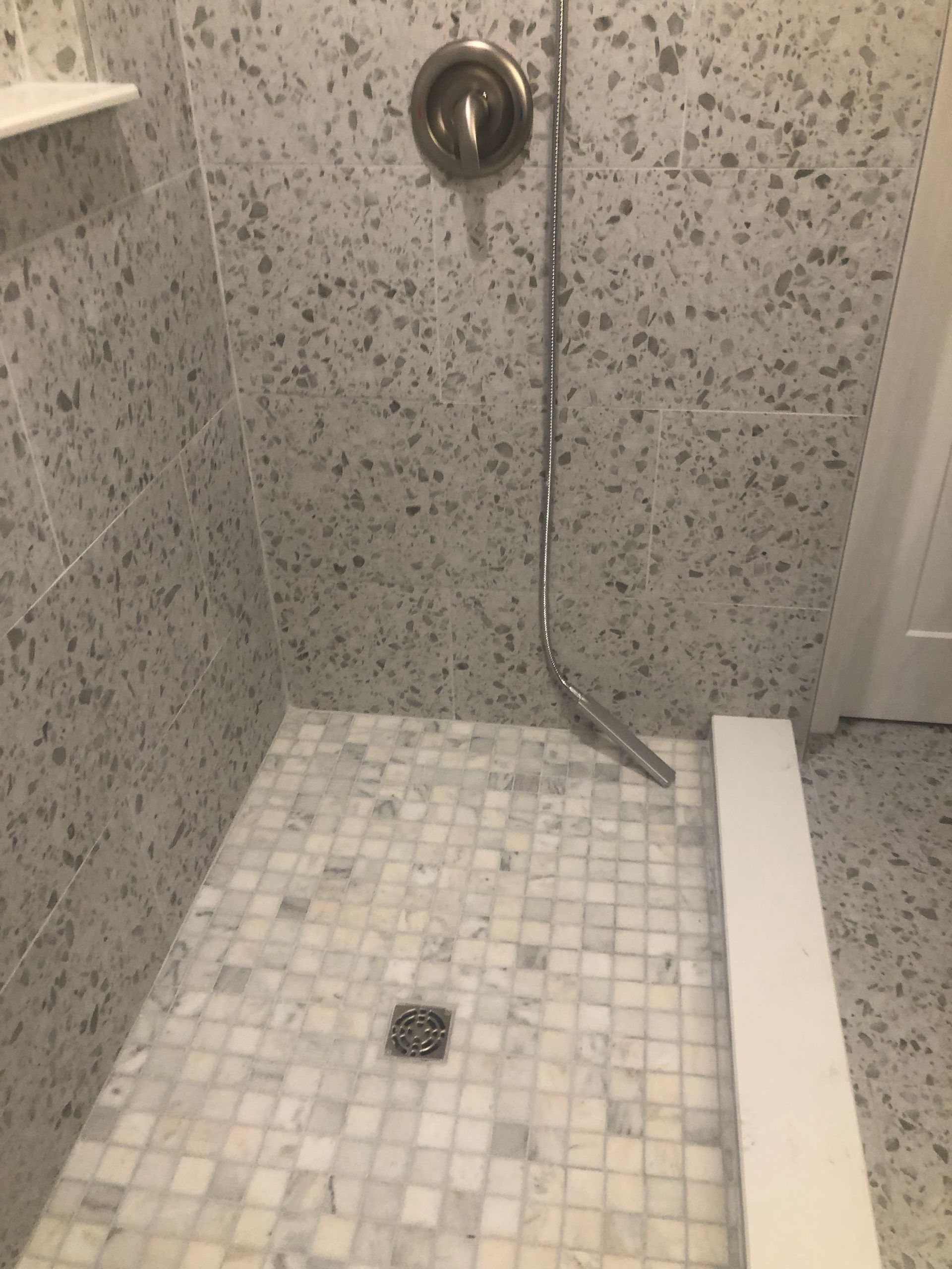 Small tiled shower stall with a wall-mounted showerhead, handheld sprayer, and center drain.