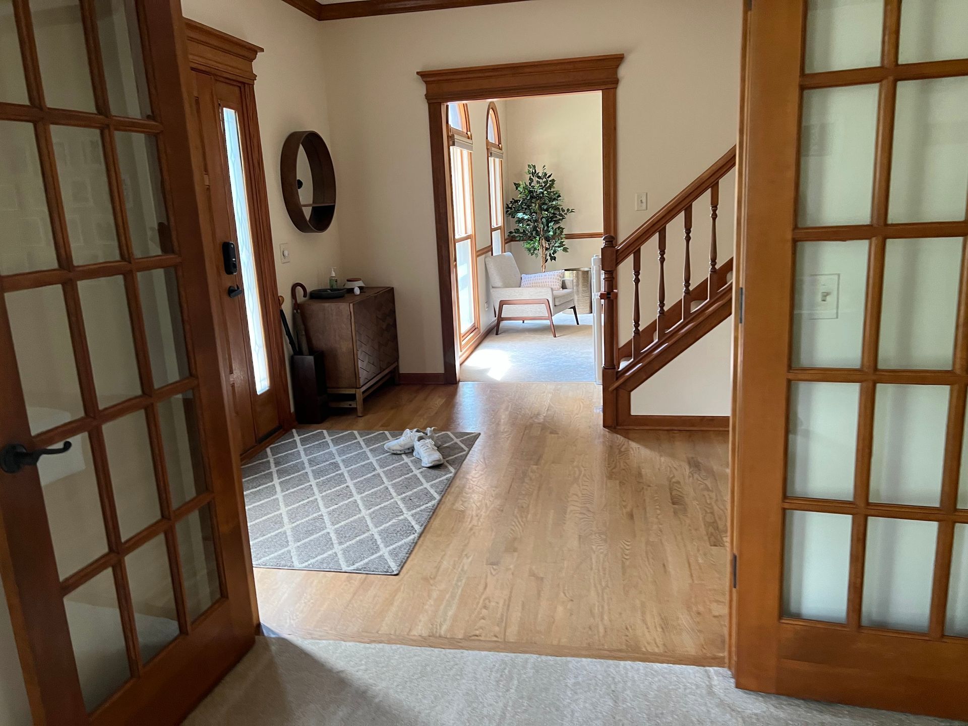 Wood-floored entry hall with glass doors, staircase, rug, and a view into a bright room beyond