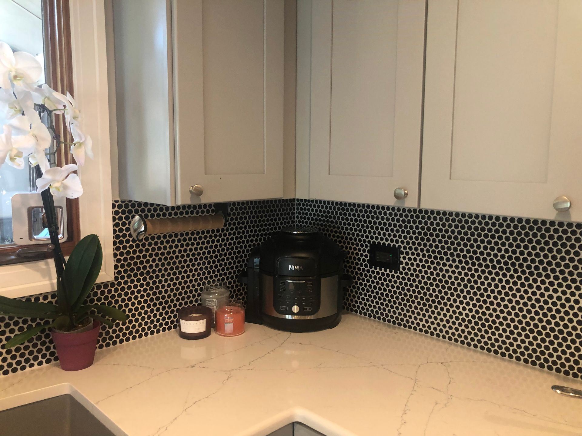 Kitchen countertop with coffee maker, black tile backsplash, white cabinets, and potted plant by the window