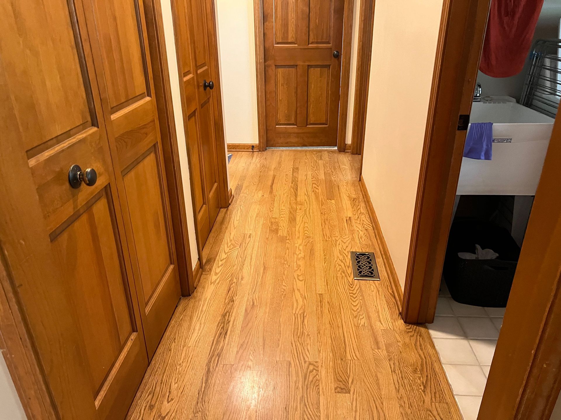Narrow wooden hallway with cabinets and doors, light wood floor, and a small floor vent.