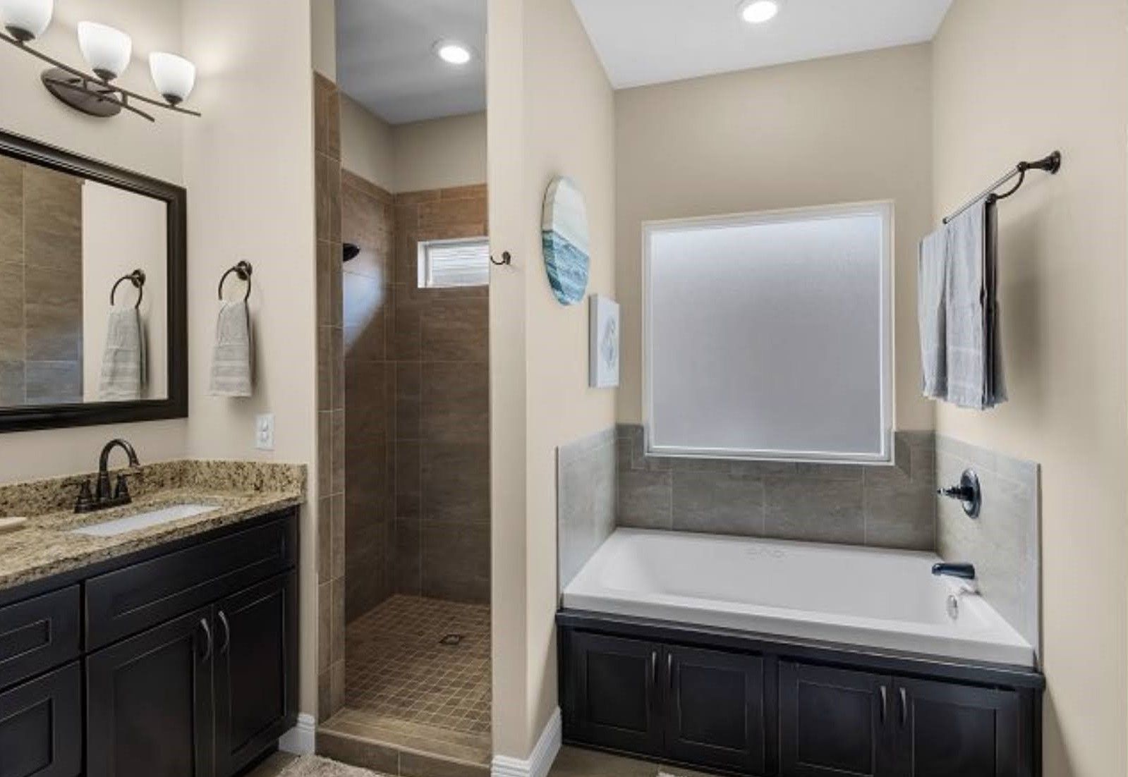 Small bathroom with dark vanity, mirror, granite countertop, walk-in shower, and a white tub beneath a frosted window
