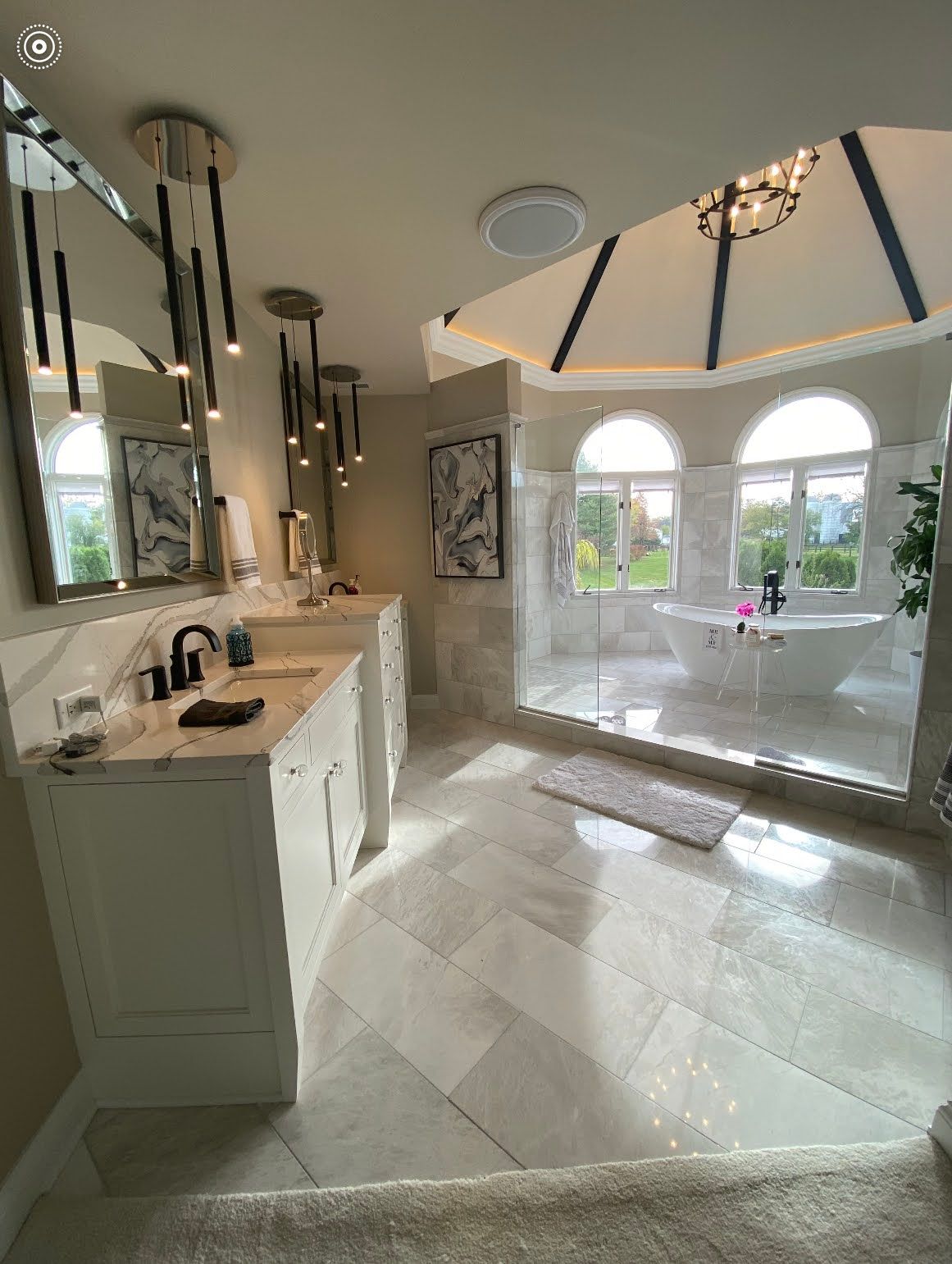 Bright modern bathroom with double vanity, stone floor, arched windows, and chandelier lighting