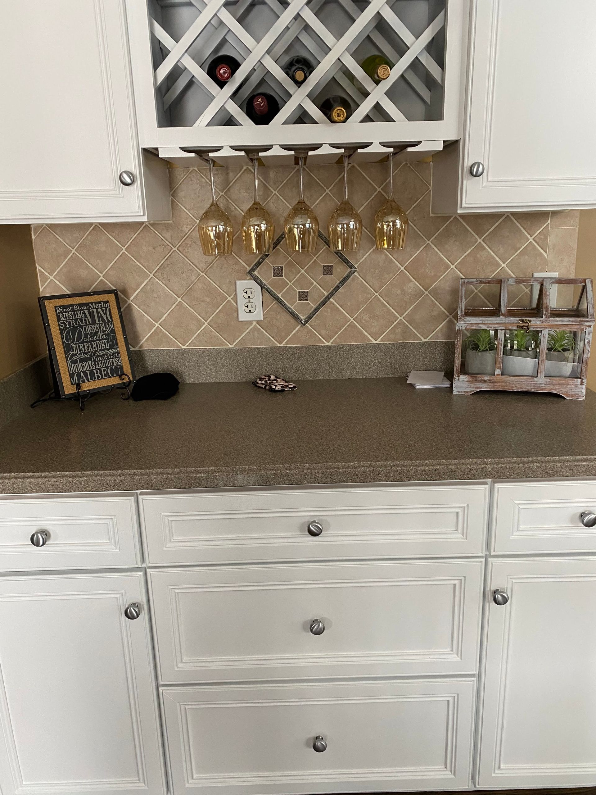 White kitchen counter with wine glass rack, granite backsplash, hanging mugs, and white cabinets
