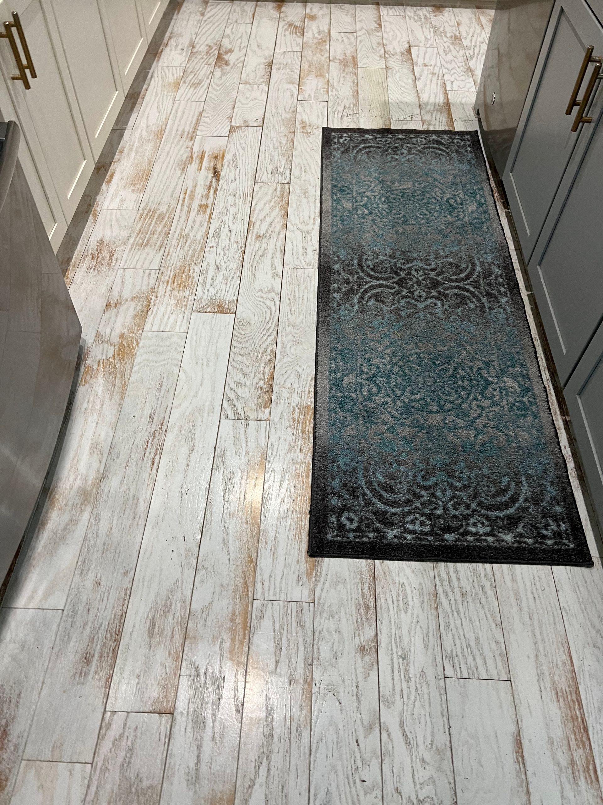Narrow kitchen hallway with white worn wood flooring and a dark rectangular rug.