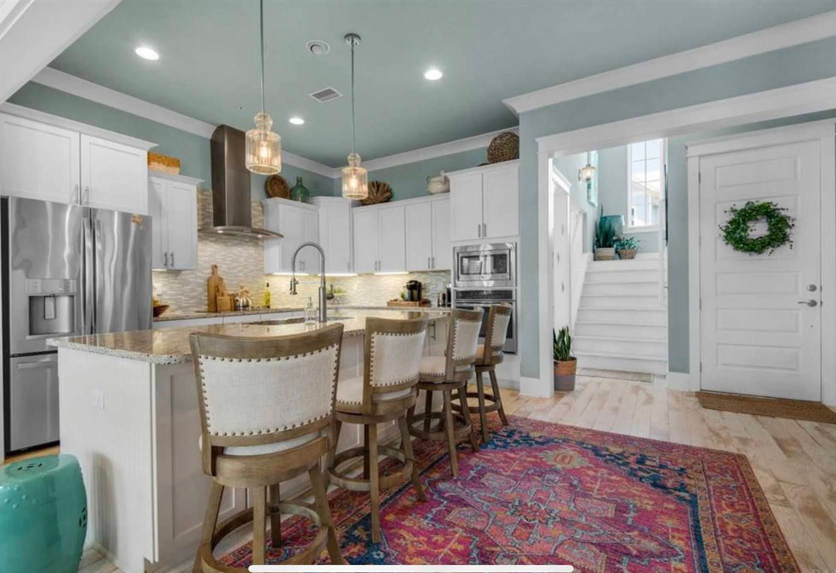 Bright white kitchen with island, barstools, pendant lights, and a colorful rug leading to a staircase.