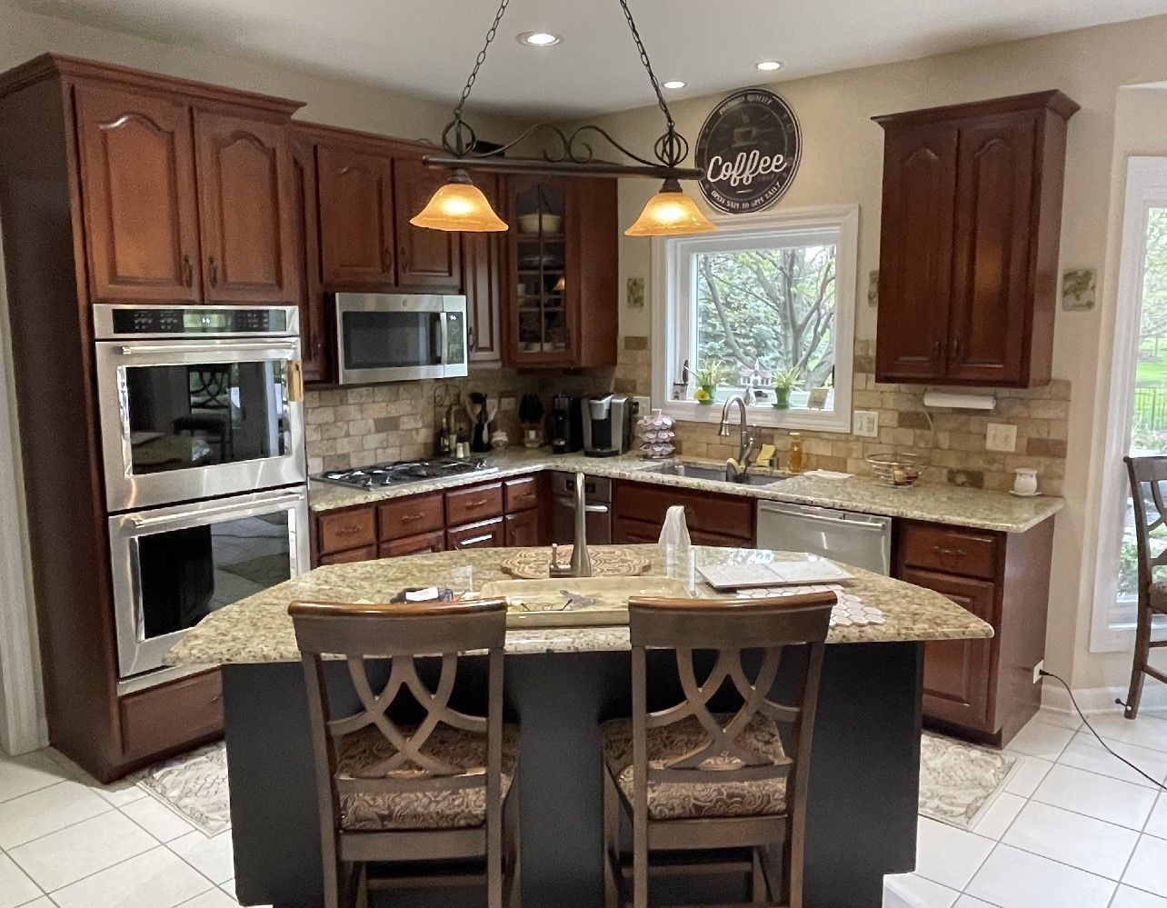 Warm kitchen with dark wood cabinets, granite island, stainless appliances, and two barstools under pendant lights
