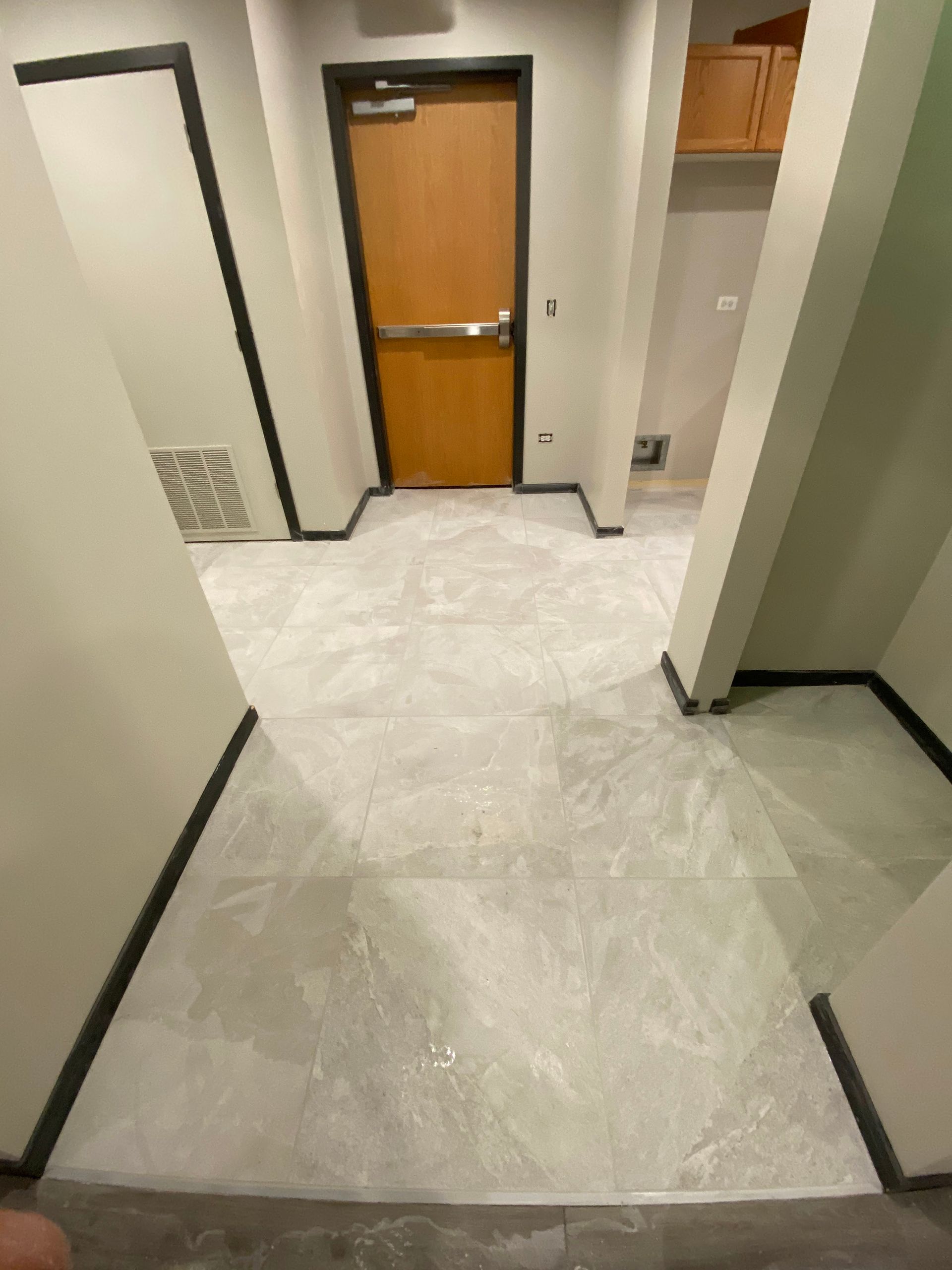 Narrow hallway with white tiled floor, beige walls, and a wooden door at the end.