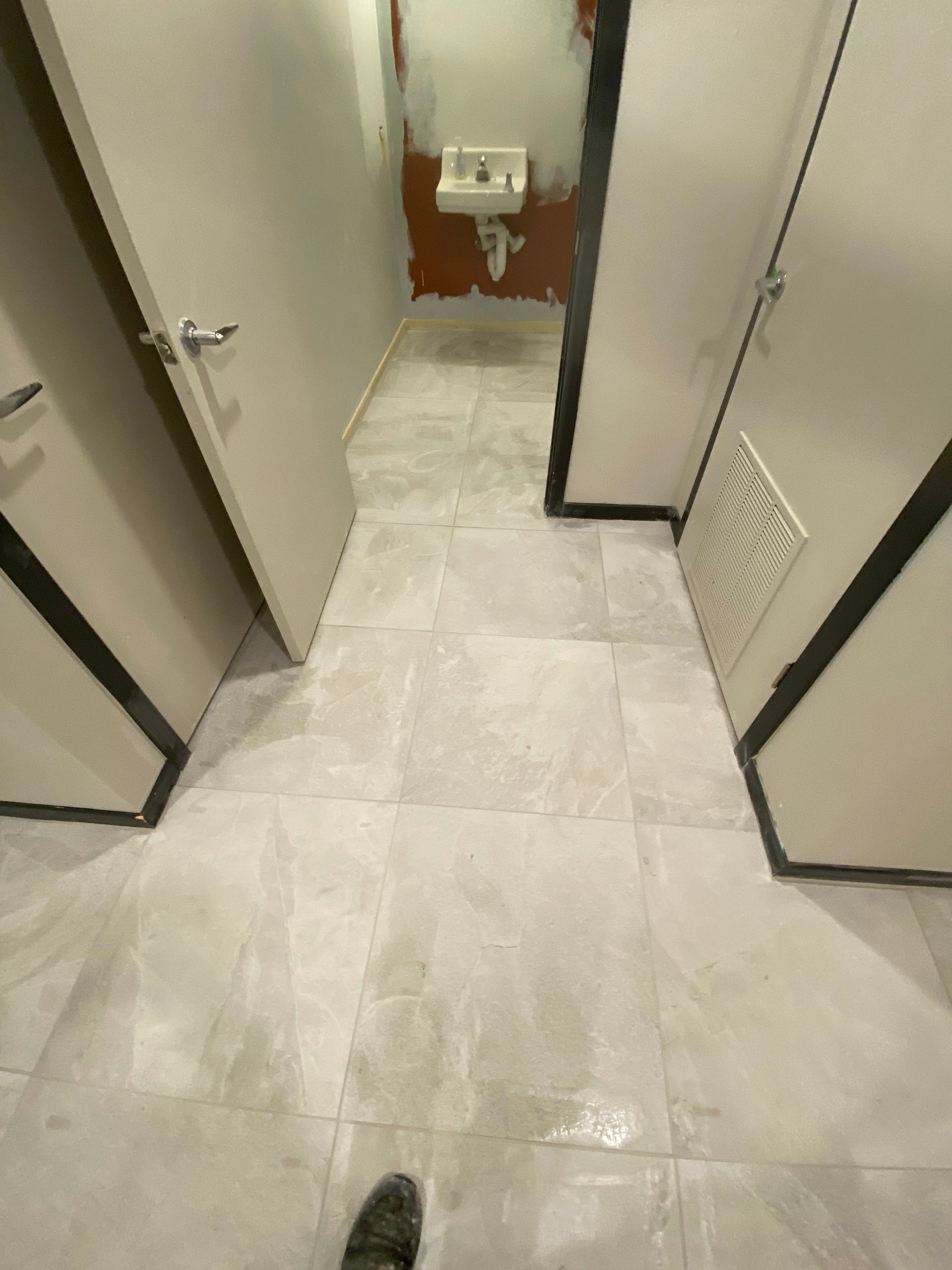 Narrow bathroom hallway with white tile floor, open stall doors, and a toilet visible at the end.