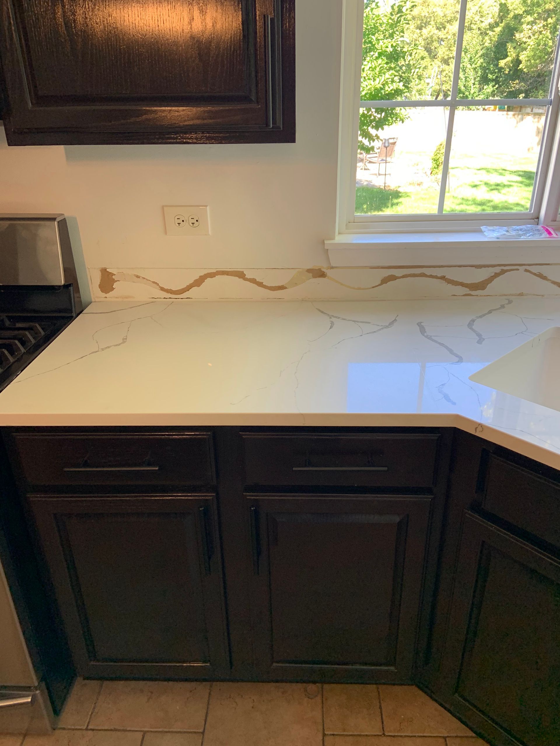 Kitchen countertop with a wavy crack along the backsplash under a window, above dark cabinets.