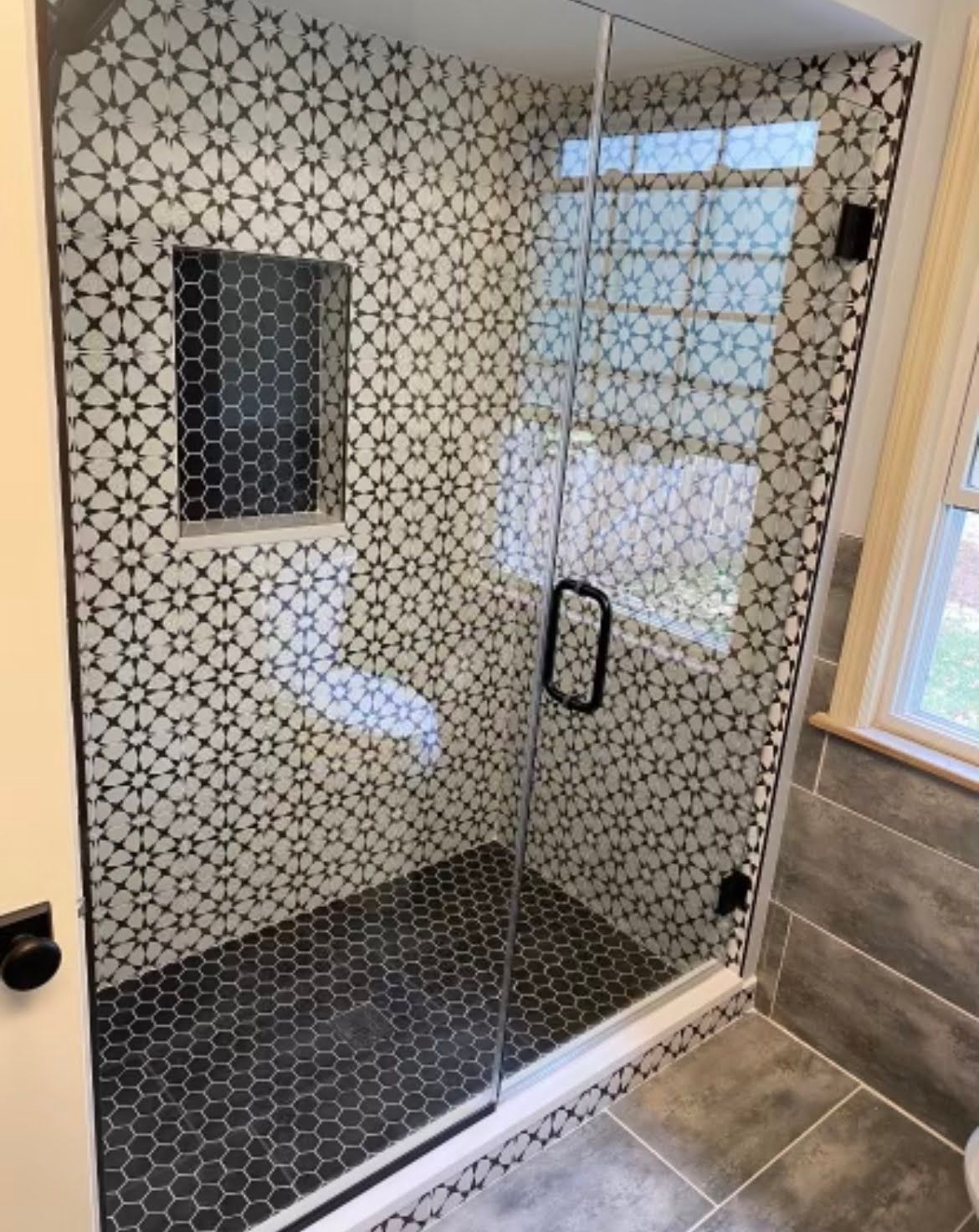 Walk-in shower with black-and-white patterned tile and glass door beside a tiled bathroom floor.