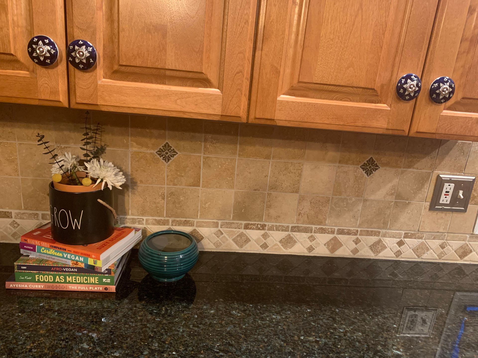 Kitchen counter with books, a small plant, teal bowl, and wooden cabinets above a tiled backsplash.