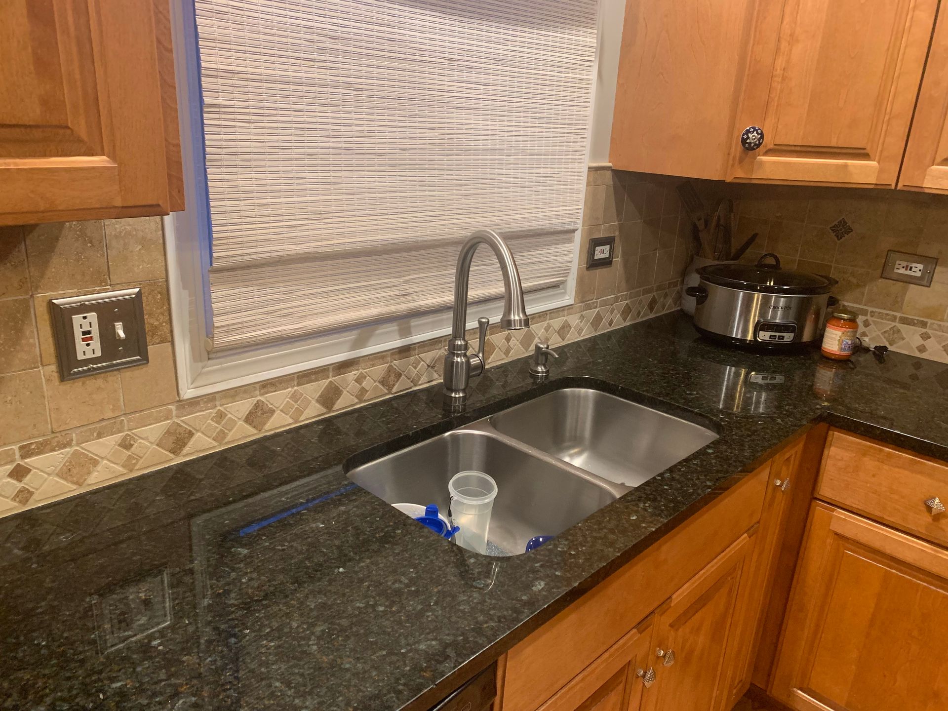 Kitchen sink with dark granite countertop, stainless faucet, and wooden cabinets under a patterned window shade