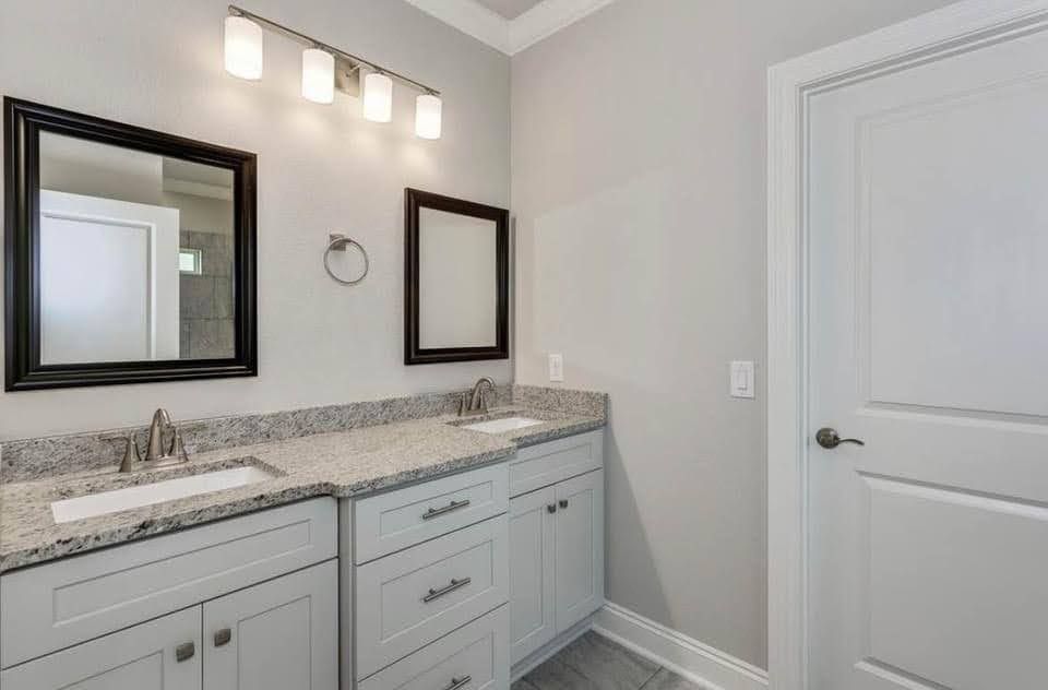 Bright bathroom vanity with granite countertop, double sinks, mirrors, and white door in a neutral gray room