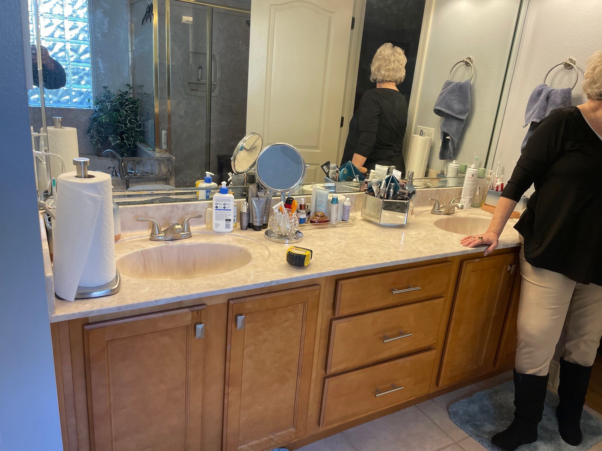 Bathroom vanity with twin sinks, mirrors, toiletries, and a person standing at the right edge