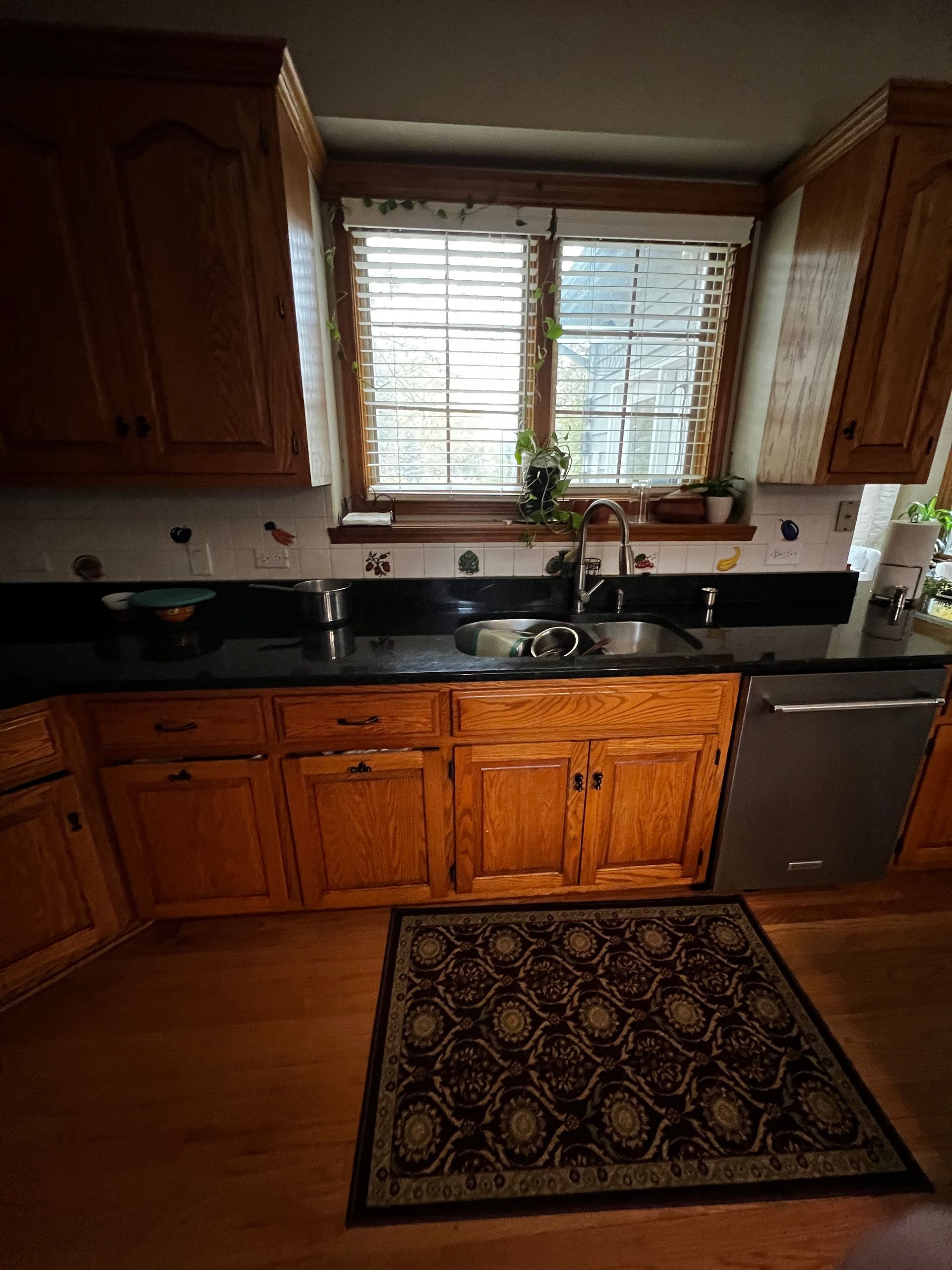 Small kitchen with wooden cabinets, black countertops, window over sink, and a patterned rug.