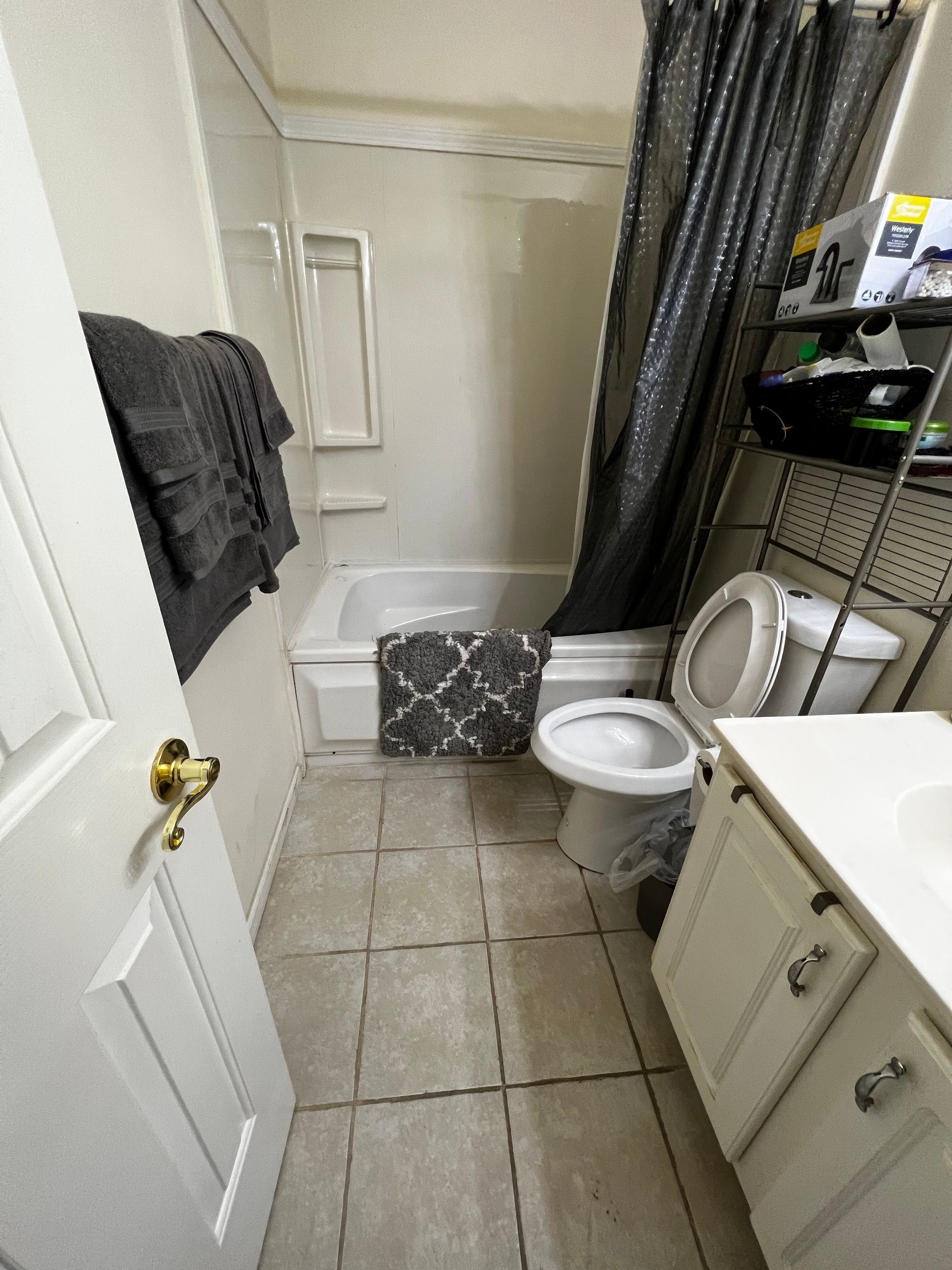 Small bathroom with white toilet, shower curtain, tiled floor, and a laundry basket near the tub.