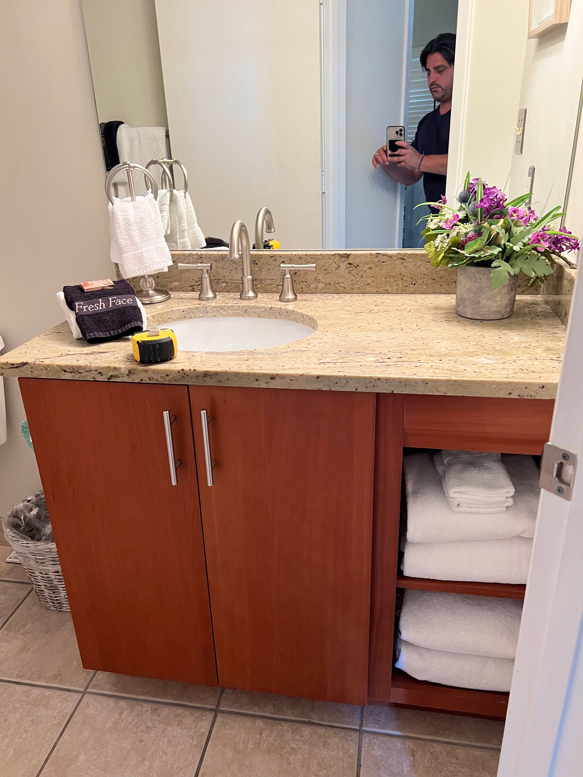 Bathroom vanity with tan countertop, wooden cabinets, mirror, and towels stacked in an open shelf