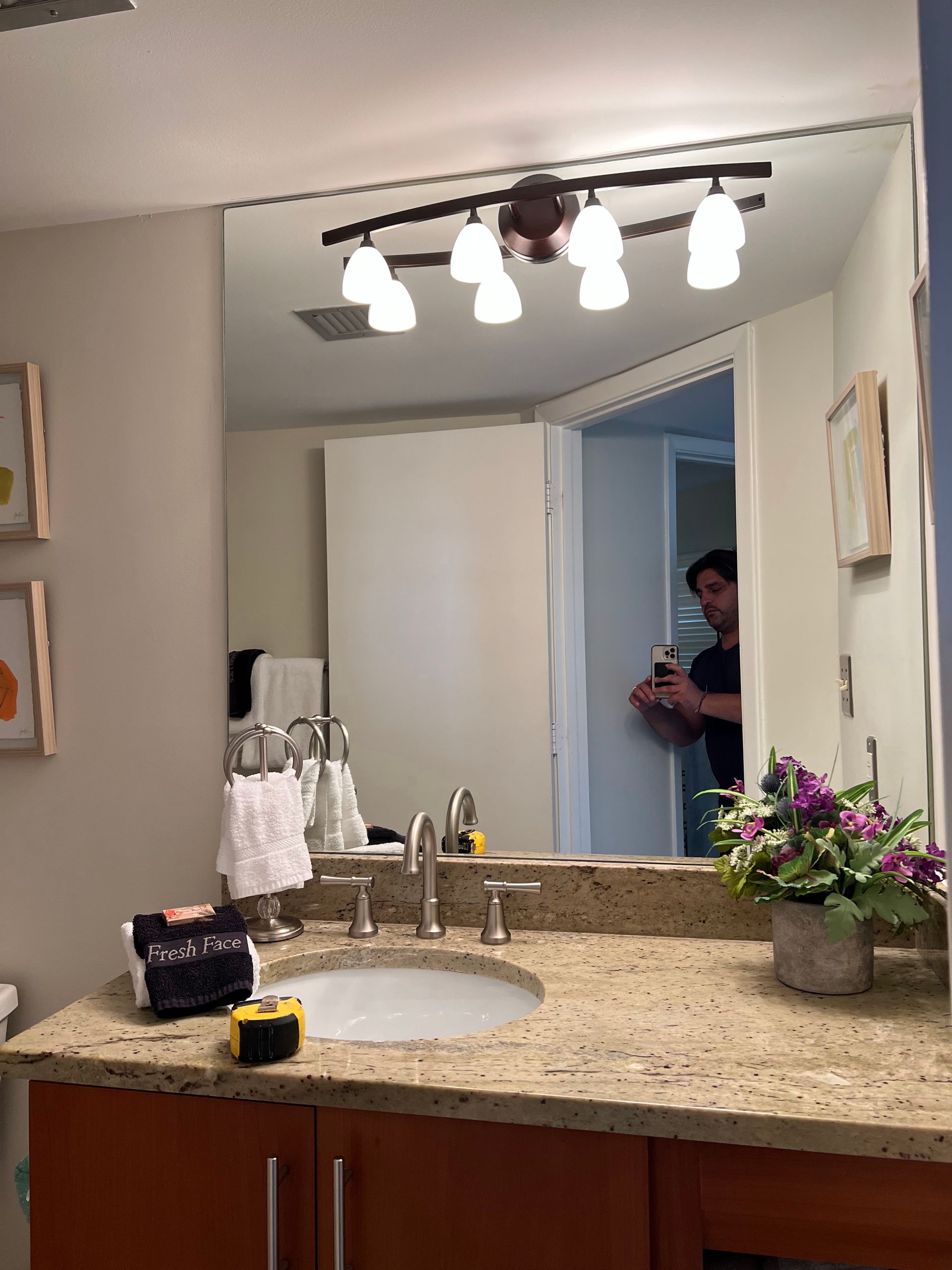 Bathroom vanity and mirror with lights, showing a person in the doorway holding a phone.