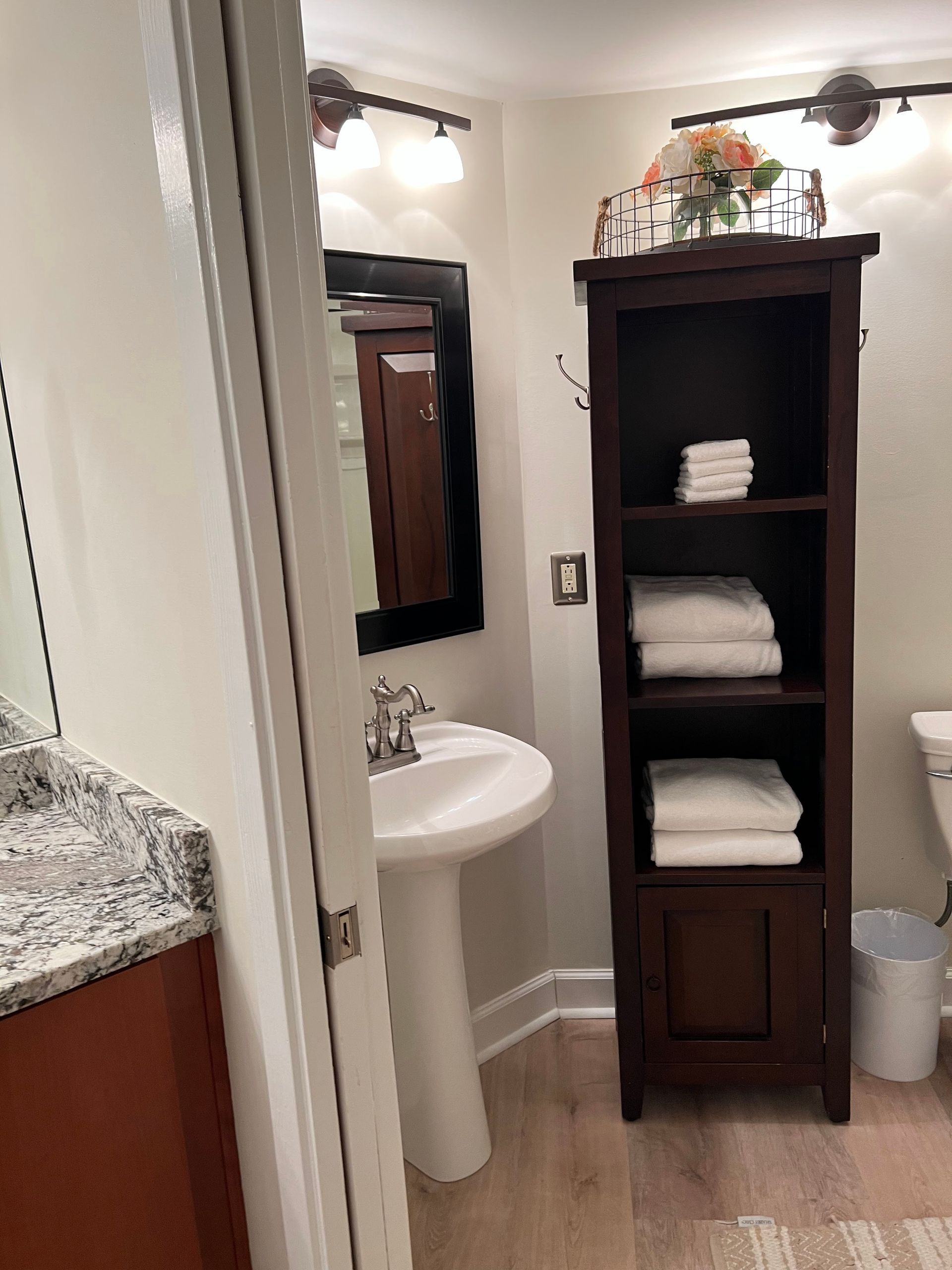 Small bathroom with pedestal sink, mirror, dark cabinet with folded towels, and granite countertop.