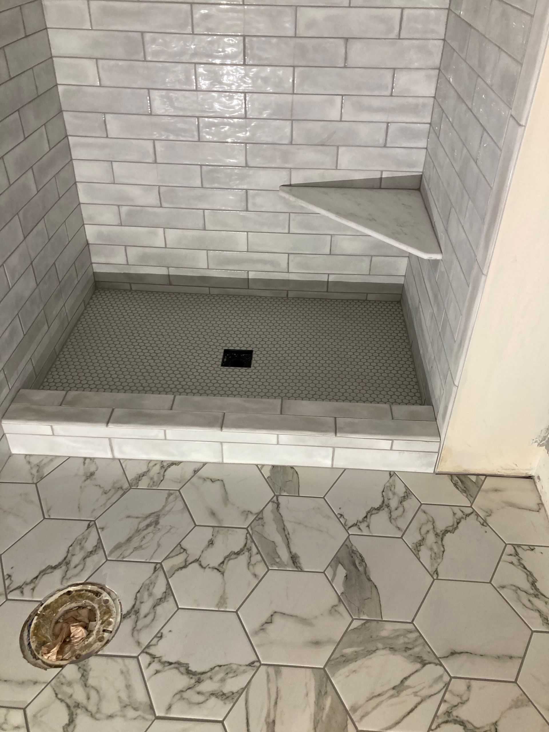 Tiled shower stall with a built-in corner shelf and floor drain, viewed above a patterned bathroom floor.