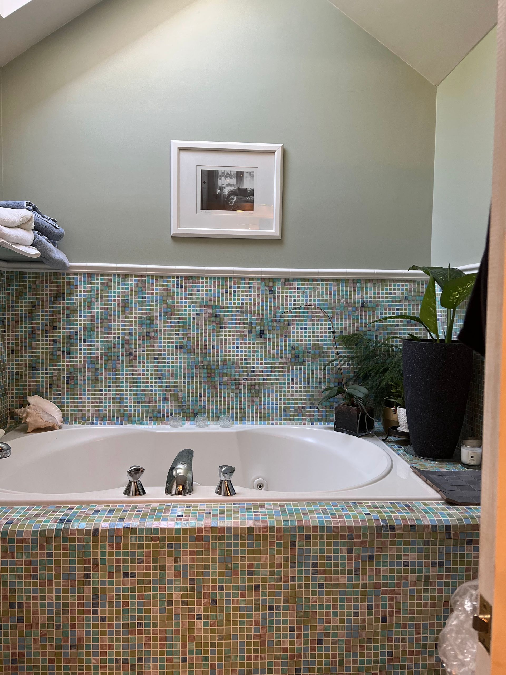 Mosaic-tiled bathroom with a white soaking tub, framed art above, and a potted plant beside it