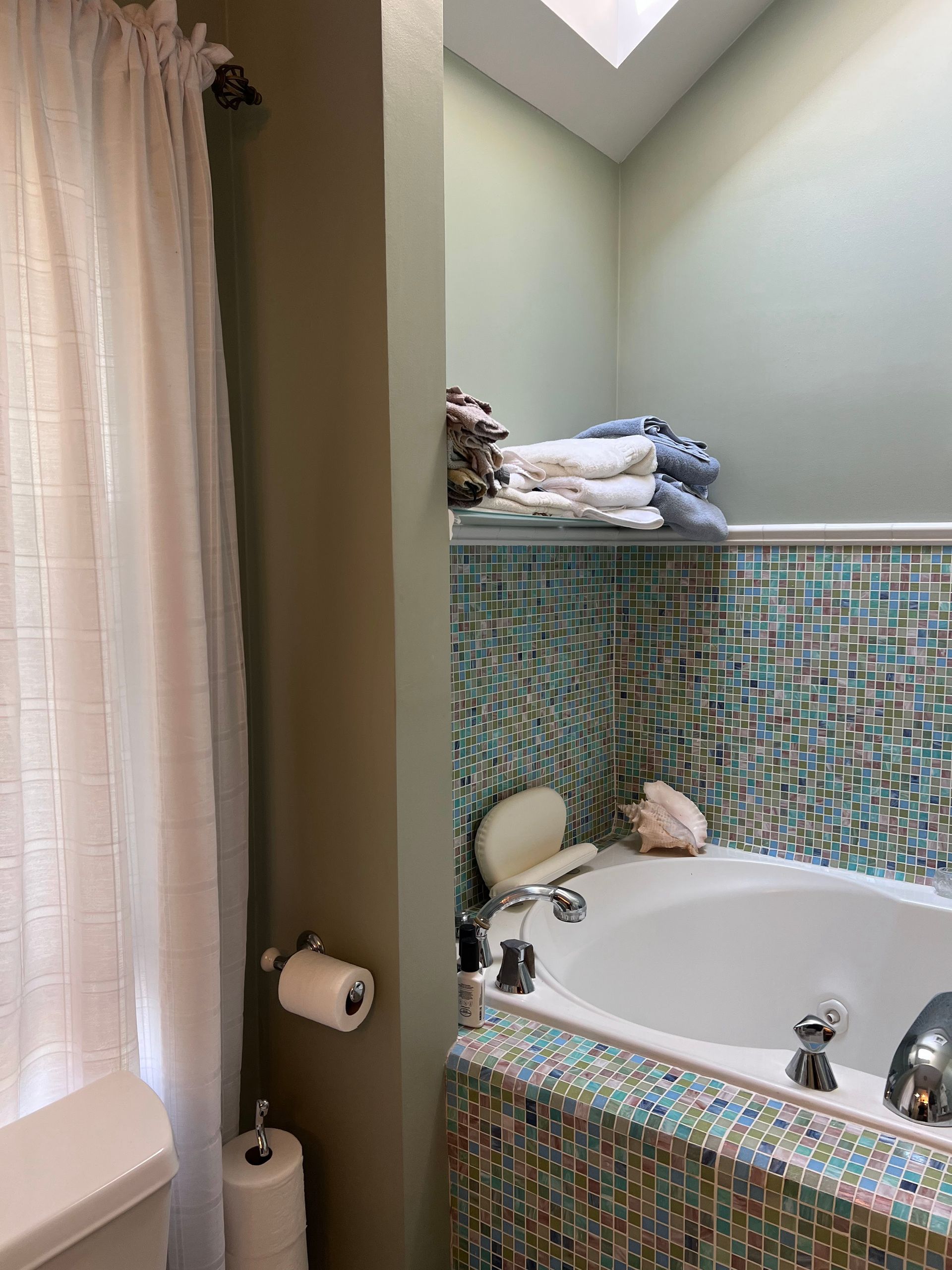 Bathroom with mosaic-tiled bathtub, toiletries on the ledge, and a window with sheer curtains.