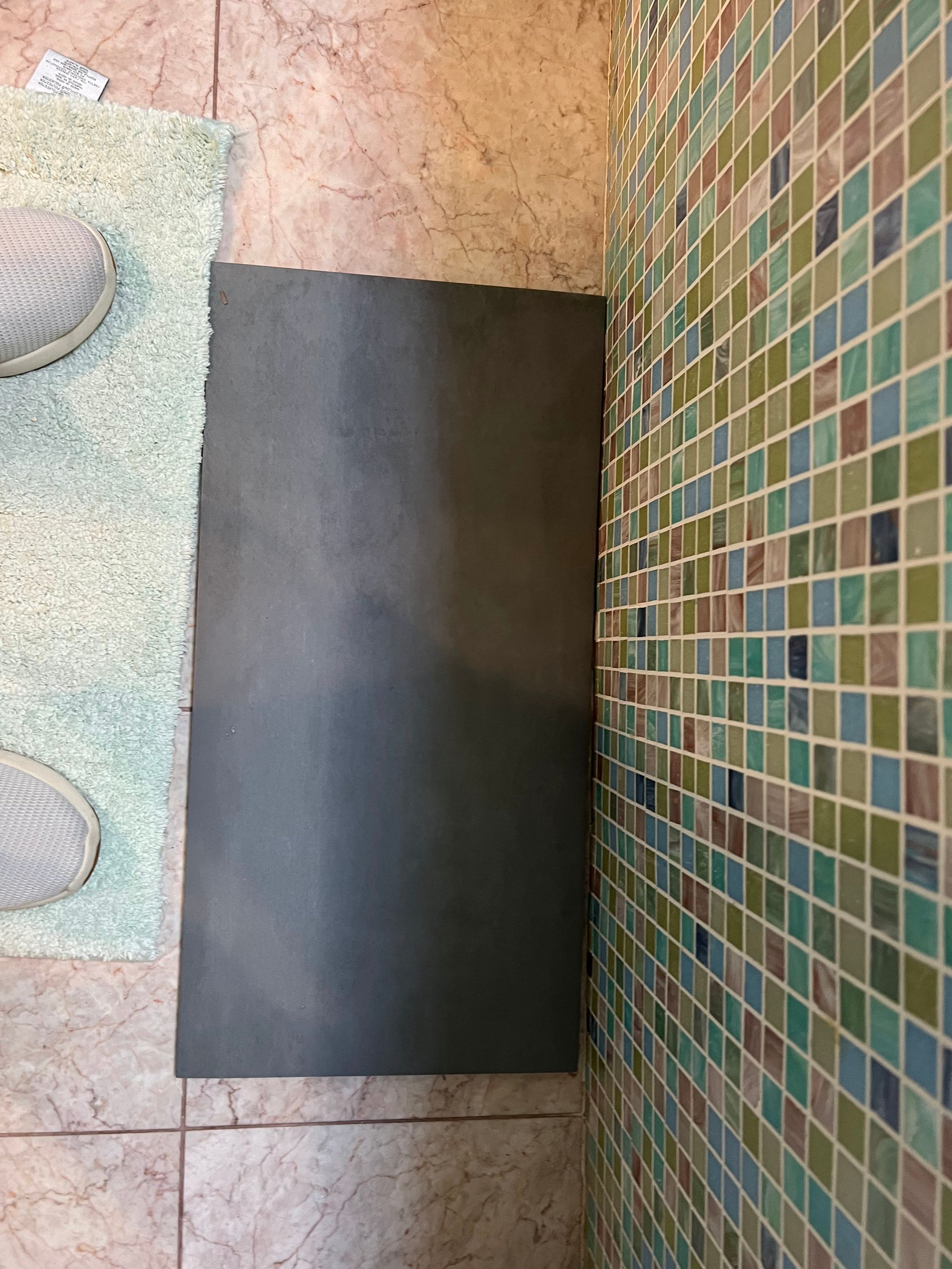 Black rectangular mat on a tiled floor beside a mosaic wall, with shoes visible at the left edge