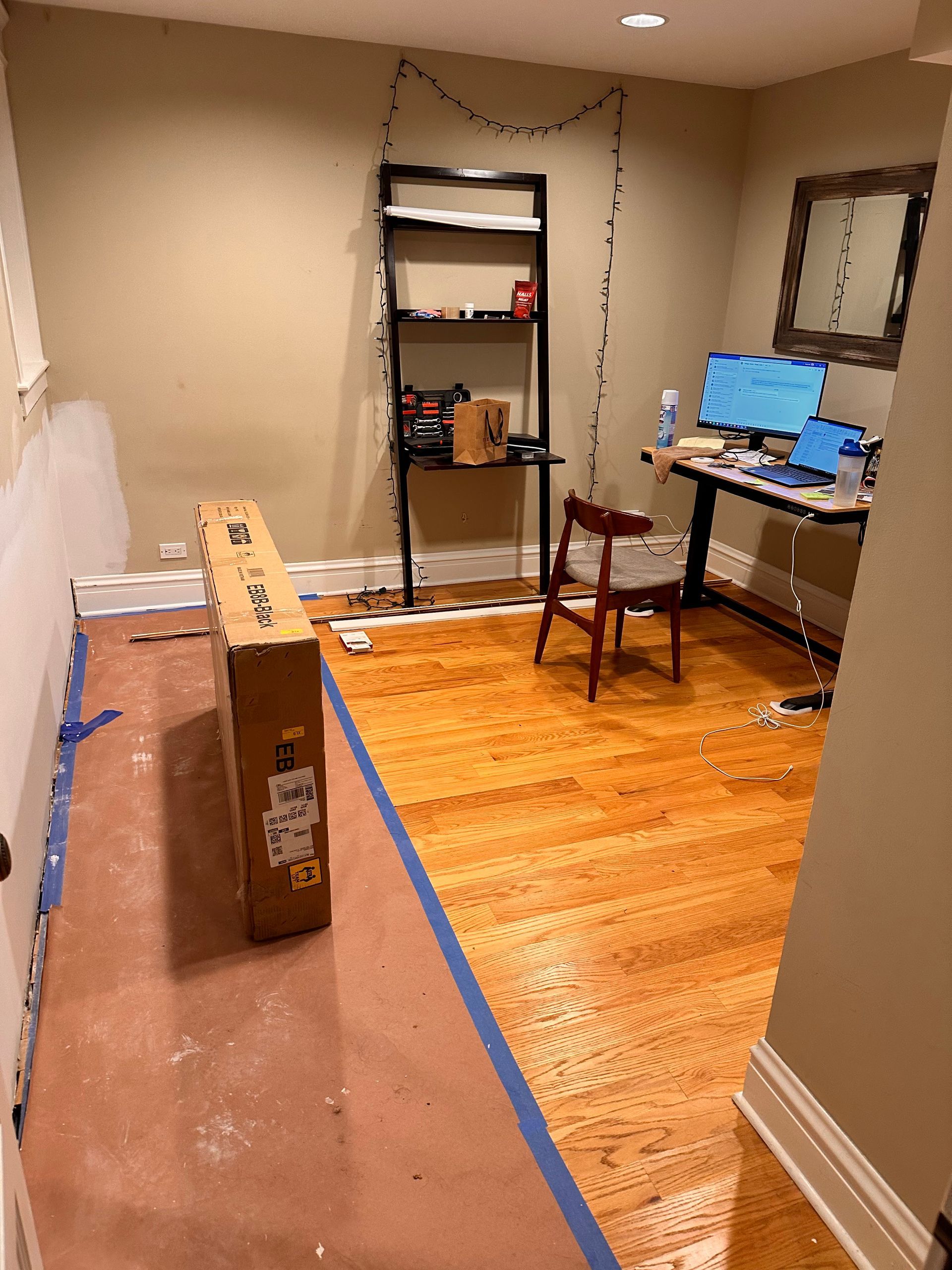 Empty room with hardwood floor, boxed item on the left, and a desk with monitors in the back corner.