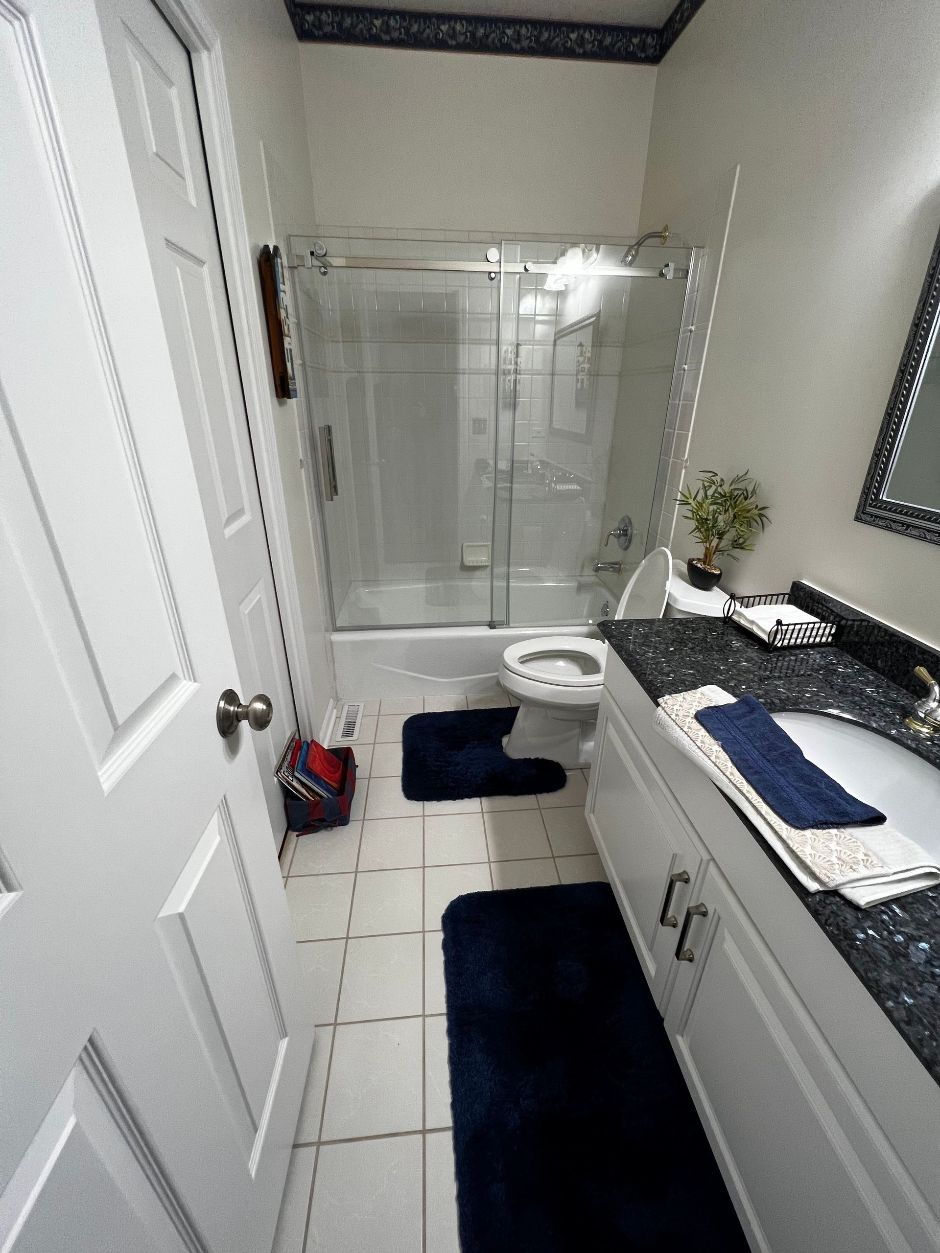 Narrow bathroom with white tile, glass shower, dark rugs, and a vanity with a blue towel.