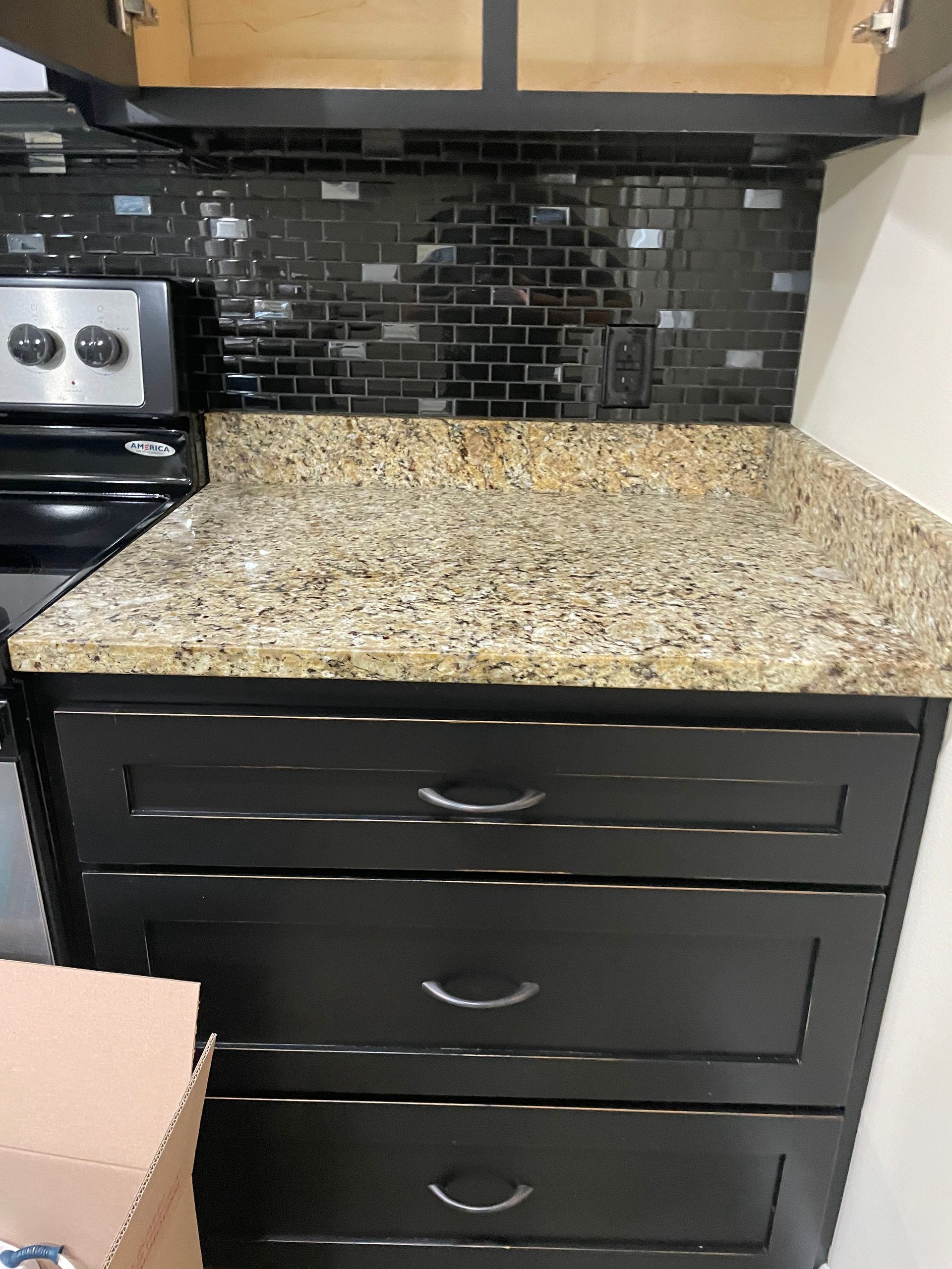Kitchen countertop and black drawers with granite top, dark tile backsplash, and stove at left.