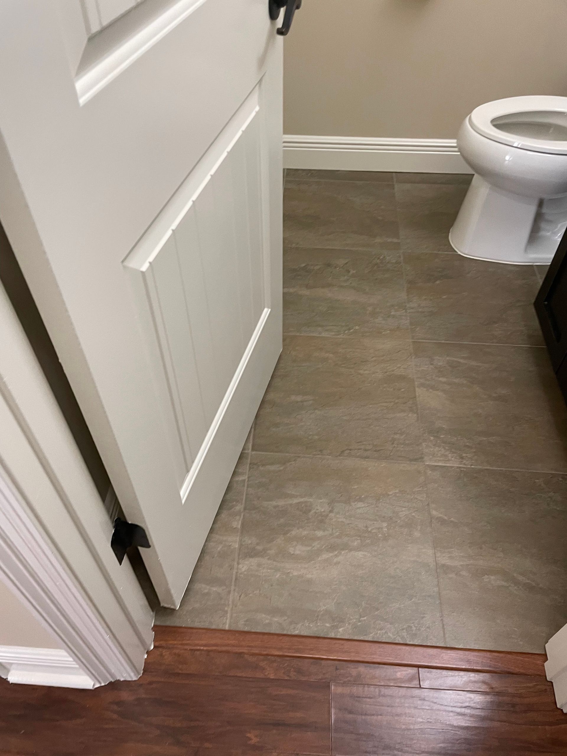 Open bathroom door with tile floor and a toilet visible inside, beside a wood hallway floor